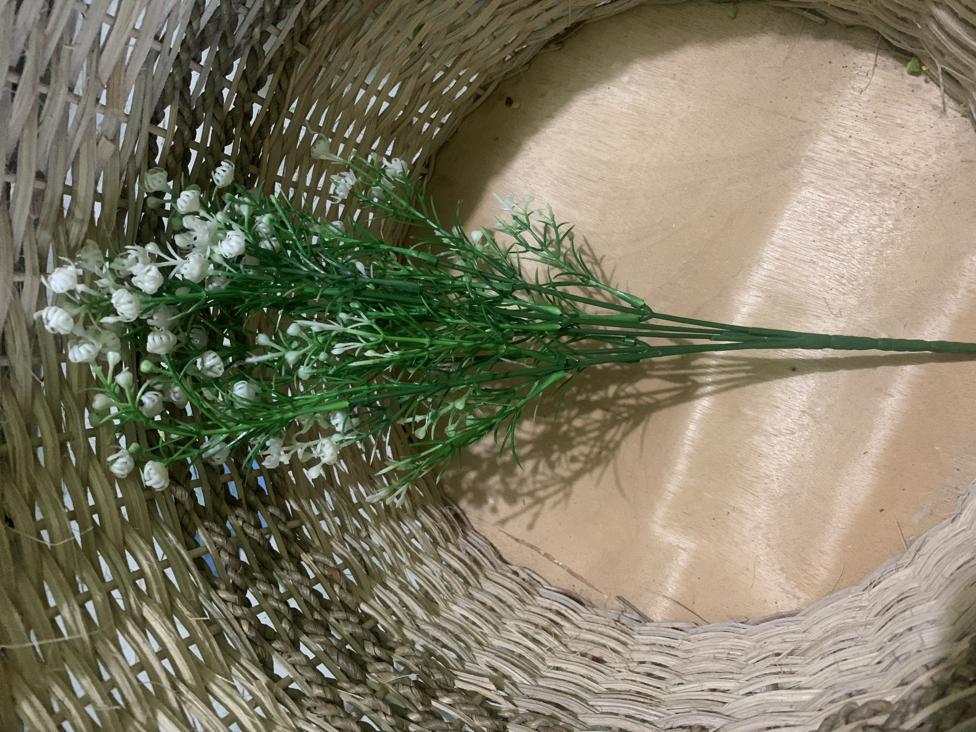 The image shows a bunch of artificial white baby's breath flowers with green stems, placed inside a round, woven basket. The basket is made of a light brown, natural material and has a distinctive weave pattern with visible gaps. The interior of the basket reveals a light brown, wooden surface. The lighting suggests an indoor setting, casting a shadow of the flowers onto the wooden surface. No people or animals are visible. The scene is static and appears to be a still life arrangement. The location in Kuala Pembuang, Indonesia, is not discernable from the visual information provided in the image.