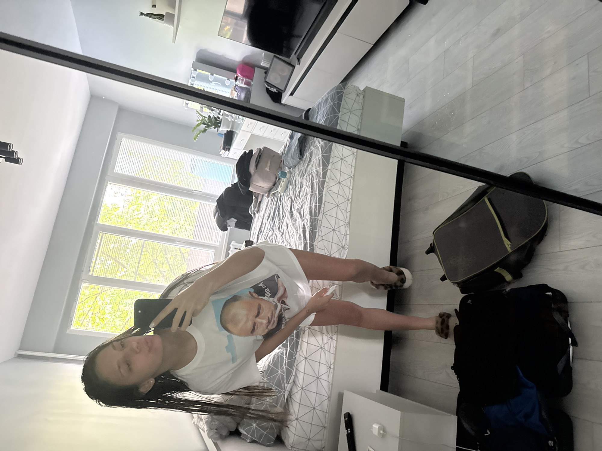 A young woman is taking a selfie in a mirror in a bedroom. The bedroom contains a bed with grey and white patterned bedding, a TV mounted on the wall with a white cabinet beneath it, a window with blinds, luggage, and a white nightstand. The location is likely in Sofia, Bulgaria, but there is no way to confirm based solely on the image.