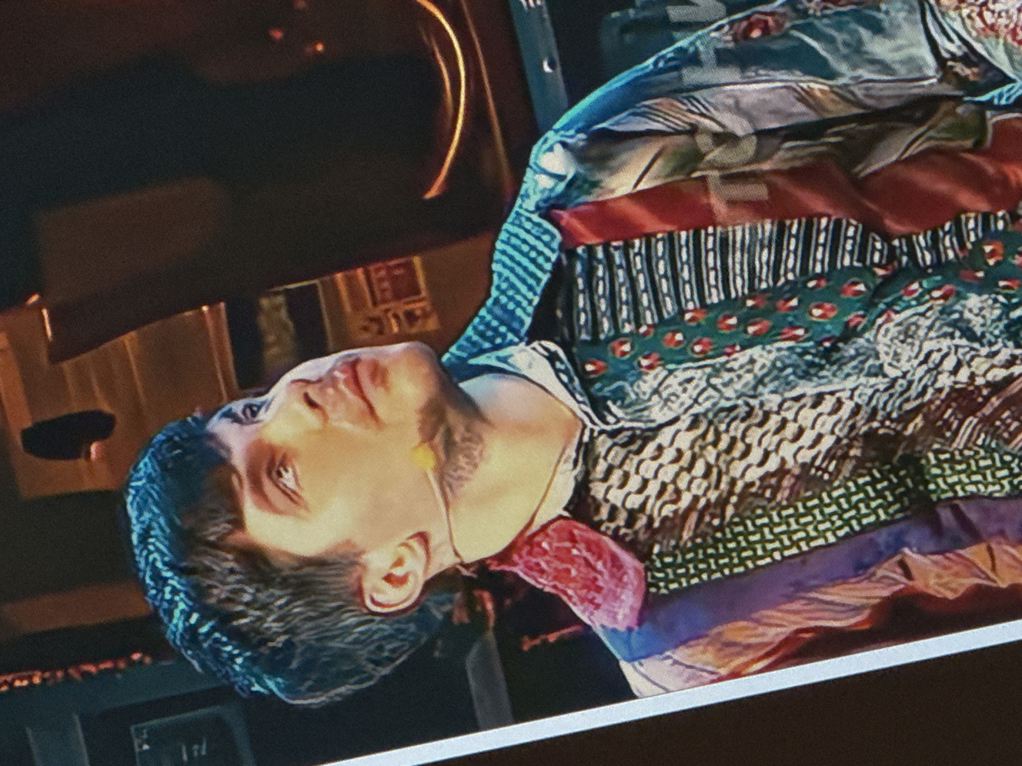 The image shows a medium shot of a young man with dark, short hair and a short beard. He is wearing a colorful, patterned shirt with various designs including stripes, geometric shapes, and what appear to be abstract images. The shirt has a prominent collar with a red, textured detail. A thin microphone wire is visible around his neck, with a small yellow microphone element near his chin. The man is looking upwards and to his right, with his mouth slightly open. The background is dark and appears to be an indoor setting, possibly a stage or studio, with some indistinct illuminated elements and structures that are not clearly identifiable. There is no visible connection to Saratov, Russia in the image.