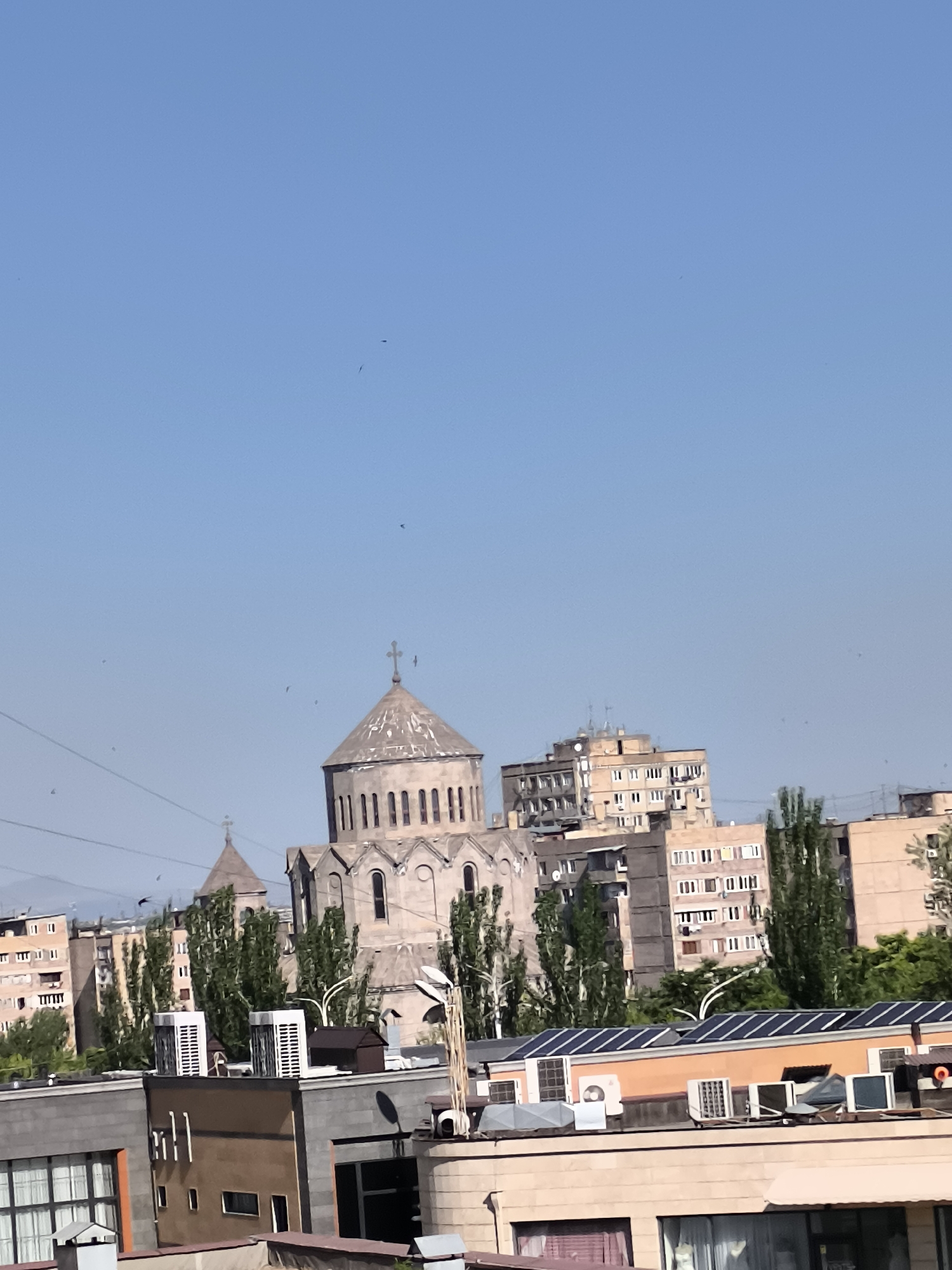 The image captures a vibrant, sunny urban scene in Yerevan, Armenia. At the center of the frame stands a prominent stone church, featuring a large central dome and a smaller one to its left, both topped with crosses, showcasing traditional Armenian architectural style.
Surrounding the church are numerous multi-story residential or commercial buildings, characterized by their blocky appearance and visible balconies. Tall, slender green trees are interspersed among the buildings, adding a touch of nature to the city environment. In the foreground, rooftops of other buildings are visible, equipped with various modern amenities such as multiple air conditioning units and arrays of solar panels, suggesting a focus on climate control and renewable energy. Power lines are also visible in different parts of the scene.
The sky is a clear, bright blue, indicating a sunny day. Several small, dark specks, likely birds or insects, are visible against the expansive sky. The lighting suggests it is mid-day, with strong sunlight illuminating the cityscape. No people are explicitly visible, and there is no discernible text within the image. The overall impression is one of a bustling city under a bright, clear sky.