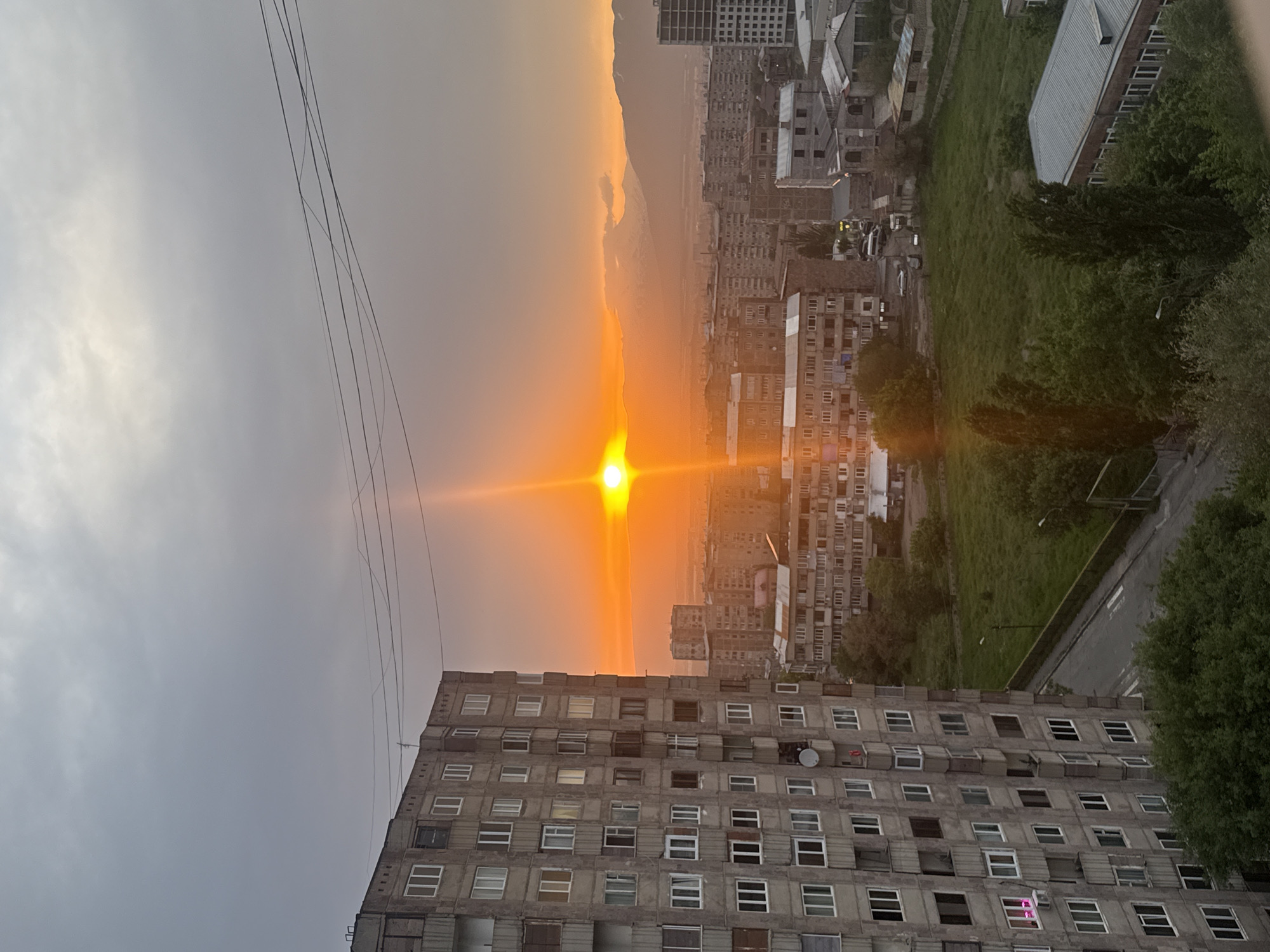 The image shows a sunset in Abovyan, Armenia, seen from a high vantage point. A large, grey concrete apartment building dominates the foreground, its rows of windows visible. Power lines stretch across the upper left. In the distance, other apartment buildings and green spaces are visible, leading to mountains on the horizon. The sun is setting directly between buildings, casting a bright orange glow across the sky and the city.