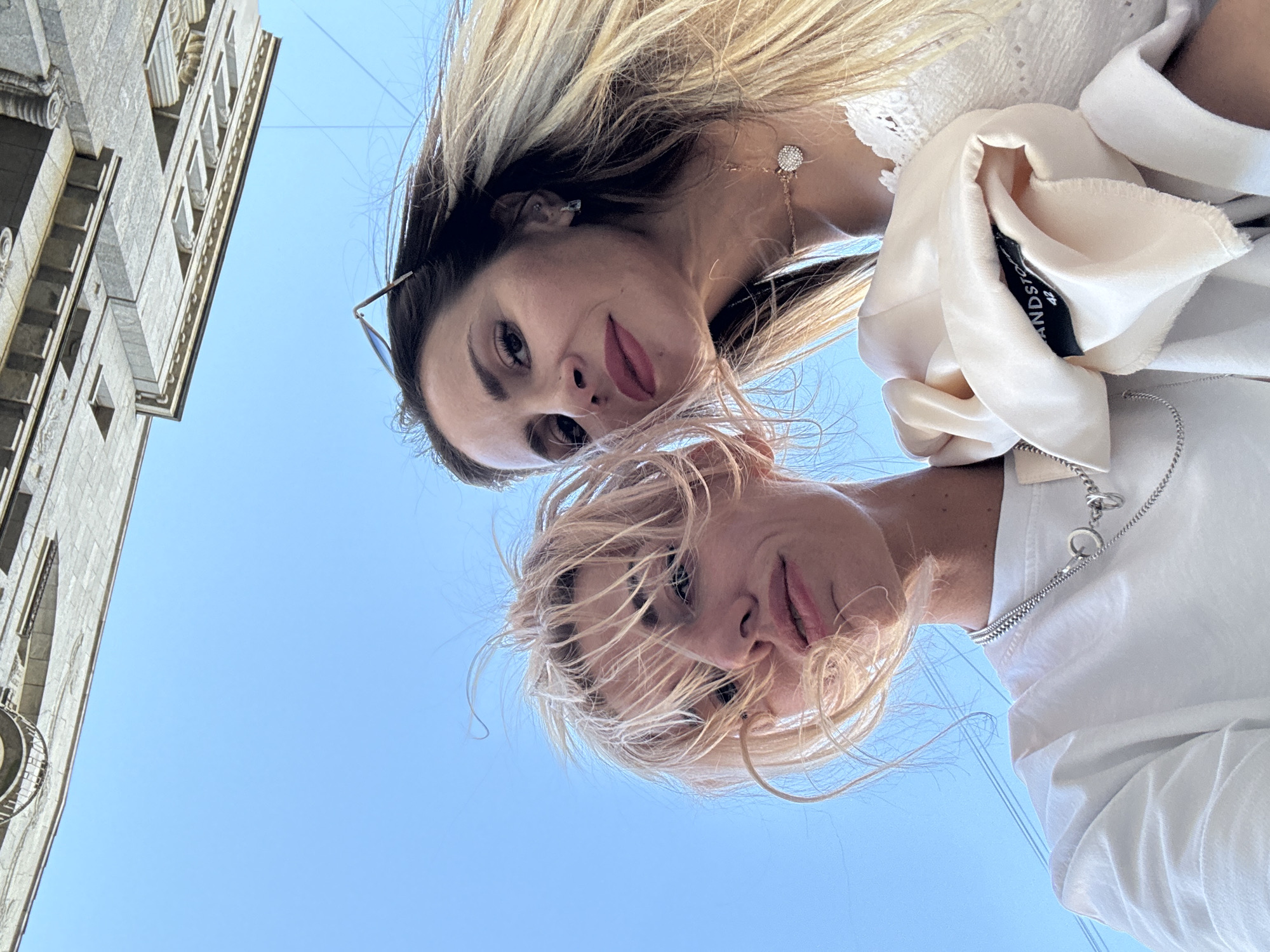 This is a selfie taken outdoors, looking up at the sky, with two women in the foreground and a building on the left. The woman in the foreground on the right has blonde hair and is wearing a white shirt. The woman to her left has darker hair, is wearing sunglasses, and has a diamond necklace. Both women appear to be smiling. Behind them, a building with architectural details can be seen. The sky is a clear blue, indicating a sunny day. The time of day appears to be daytime. There is some faint text visible on a tag attached to the white fabric in the center of the image, but it is not clearly legible. The scene suggests a casual outing in an urban environment, likely in Saint Petersburg, Russia, given the architectural style of the building and the presence of overhead wires.
