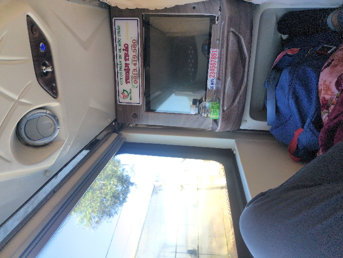 The image captures the interior of a passenger vehicle, likely a sleeper bus, viewed from a seated or reclined position. The lower-left foreground shows the bent knee and upper leg of an individual, dressed in dark gray pants. A large window on the left side of the frame provides a view of a bright outdoor environment featuring a clear blue sky, a green tree, and a light-colored building structure.
The interior ceiling above the window is light-colored and features a circular air vent and a control panel equipped with a metallic toggle switch and a blue indicator light. To the right, a dark, rectangular screen, part of a personal entertainment unit, is recessed into the pod design. Directly above this screen, a white sign with a brown frame displays Vietnamese text: "CÔNG TY CỔ PHẦN DƯỢC LIỆU THUẬN THẢO" (Thuan Thao Pharmaceutical Joint Stock Company), accompanied by a green leaf logo and the hotline number "0913.419.580".
Below the screen, a small wooden-look shelf holds a clear plastic water bottle with a green label and a white sticker marked "NK WIFI: 234567891". Beneath this shelf, an open storage compartment contains a dark blue bag with a red strap, a patterned maroon and floral blanket, and the partial view of a black shoe or foot on the far right.
The vehicle belongs to "Thuận Thảo," a company known for operating bus services that include routes to and from Dalat, Vietnam. Consequently, the scene is situated either at a bus station in Dalat or the bus is currently traversing a road within the city.