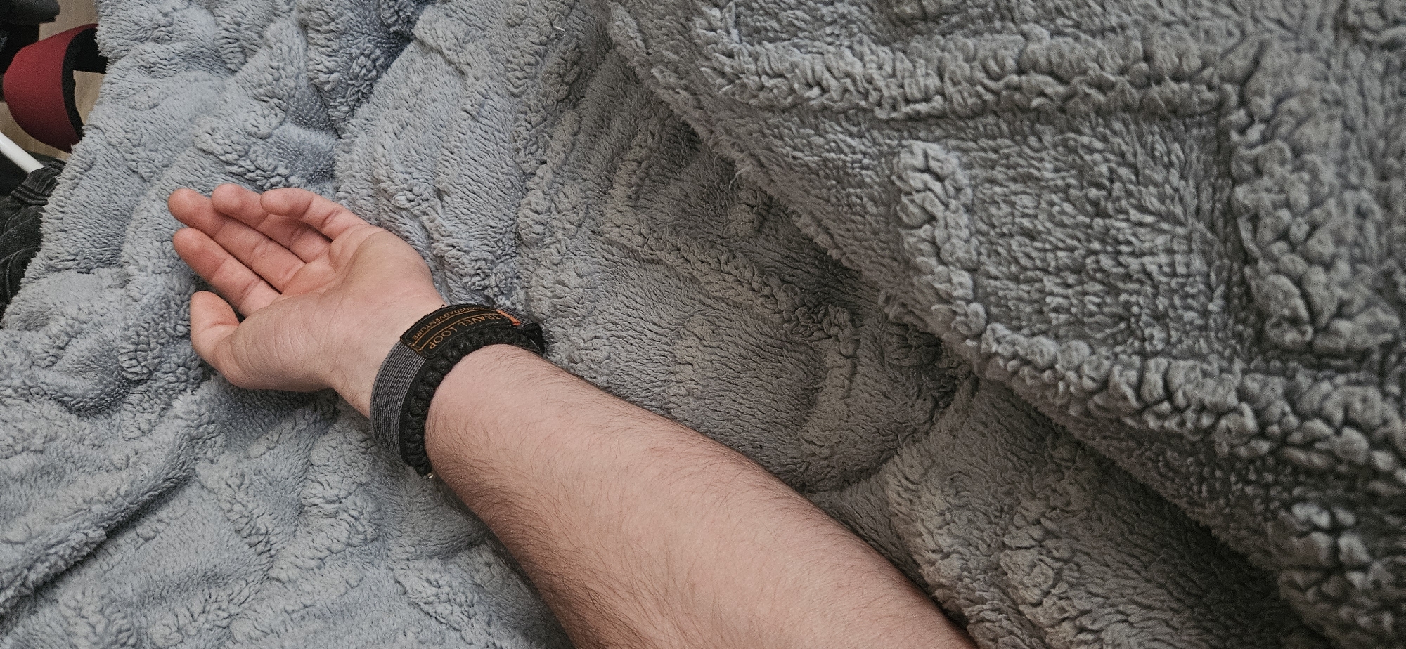 A close-up shot features a person's arm resting on a gray, plush blanket. The arm, extending from the lower left towards the upper right, is fair-skinned with visible hair. A dark bracelet with the word "Adventure" on its band encircles the wrist. The hand is open, displaying a natural skin tone. The soft texture of the blanket creates a cozy backdrop. There is no indication as to the location, Plovdiv, Bulgaria.