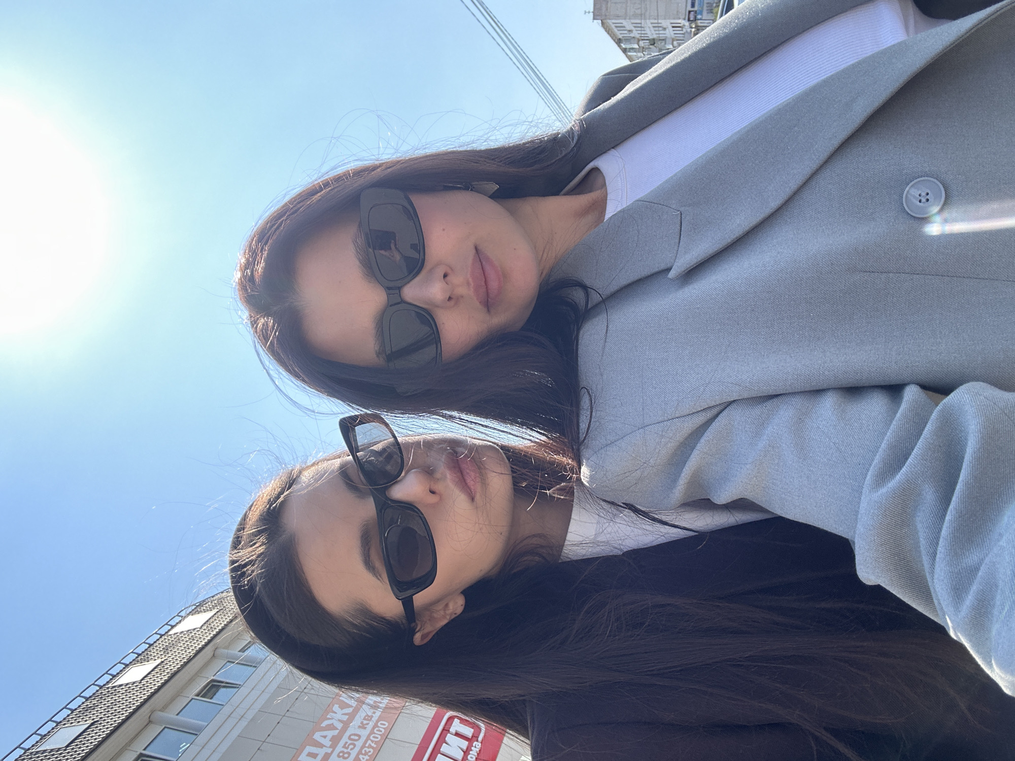 Two women are posing for a selfie outdoors. The top woman has long brown hair, sunglasses, a white top, and a gray blazer. The bottom woman has long brown hair, sunglasses, and appears to be wearing a dark jacket. They are close together. In the background, there is a building with a sign in Russian, indicating they may be in Chaikovsky, Russia. The sky is blue.
