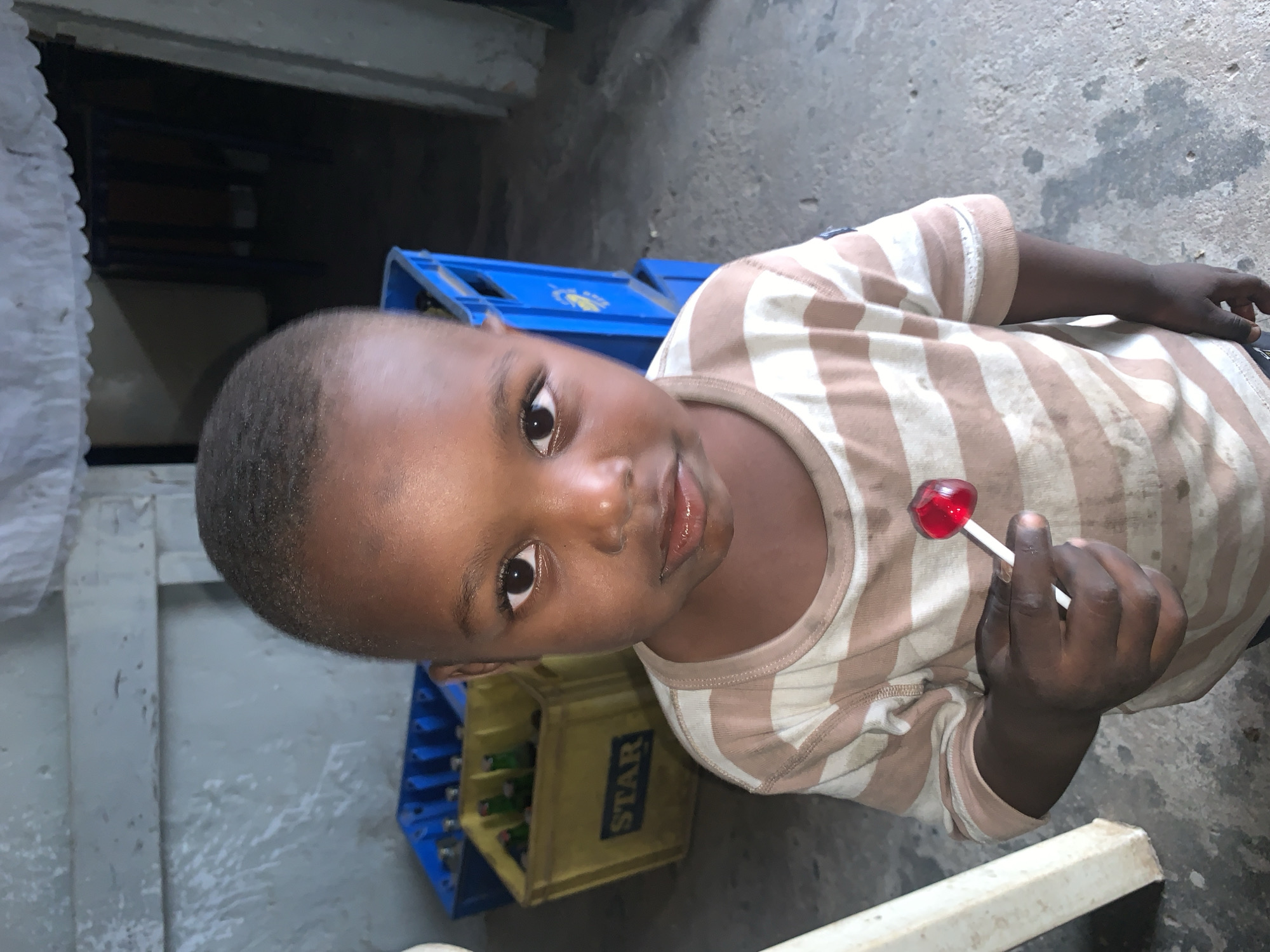 A young child, likely a boy, is the primary subject of this portrait-style image. He has dark skin, short, closely-cropped hair, and large, dark eyes that gaze directly at the viewer. His expression is neutral to slightly curious, with his lips slightly parted. He is wearing a light brown and off-white striped short-sleeved t-shirt, which appears somewhat soiled.
The child is holding a red, heart-shaped lollipop with a white stick in his right hand, positioned towards the bottom right of the frame. The background is somewhat indistinct but suggests an indoor or shaded outdoor setting with a concrete floor. To the left and behind the child, several crates are visible. One crate is bright blue, and another, yellow, has the word "STAR" printed on its side, partially obscured. These crates appear to be beverage crates, possibly for bottles, indicating a commercial or storage area like a shop or backyard. The lighting appears to be natural, possibly from an overhead source or an open doorway, casting soft shadows. The overall atmosphere is simple and everyday, capturing a candid moment of the child. The image is rotated 90 degrees clockwise, making the child's head appear to be on the left side of the frame with his body extending towards the right.