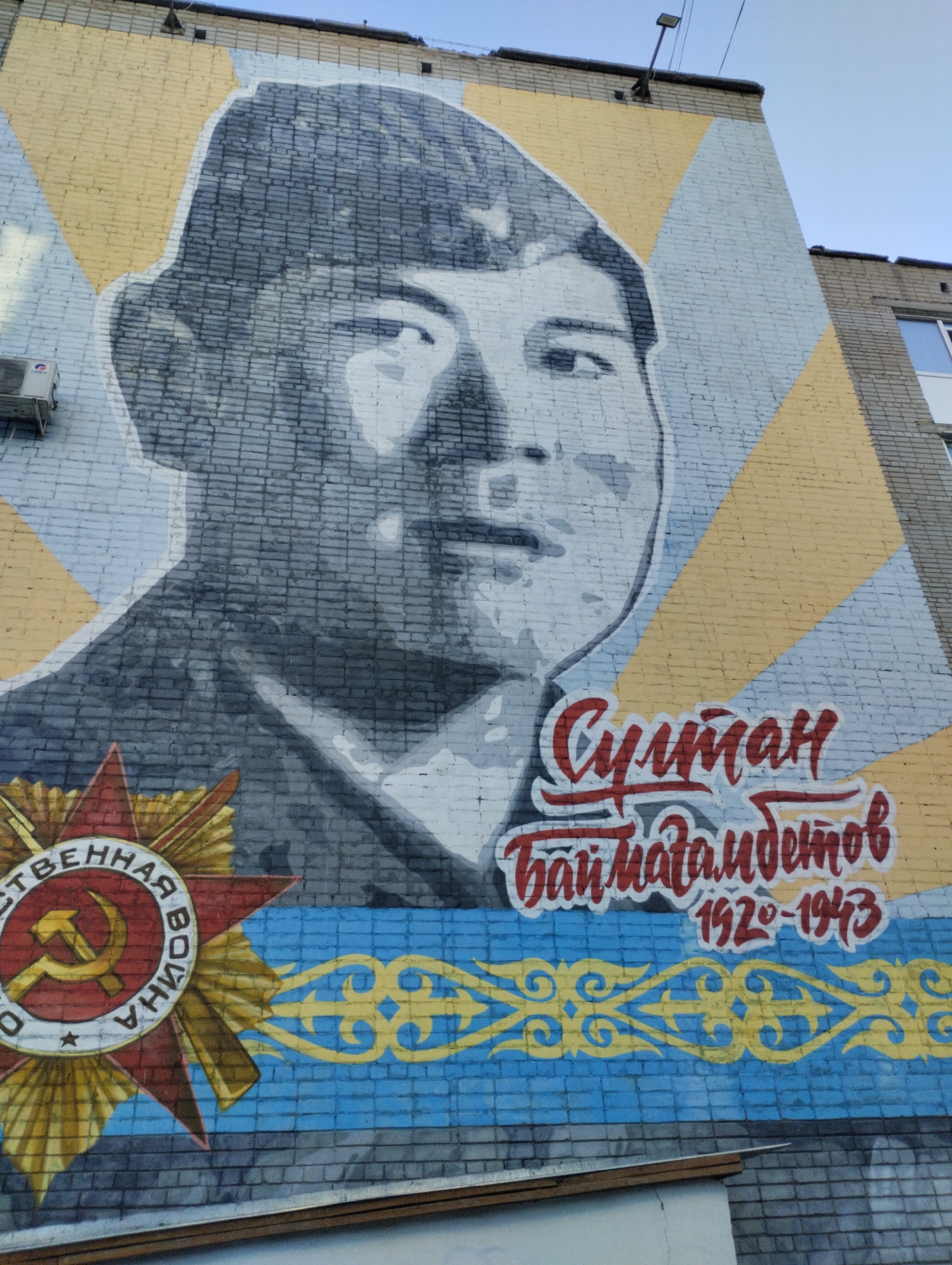 A mural painted on the side of a brick building depicts a man's portrait. The man is wearing a military-style cap and is rendered in grayscale. The background is a pattern of yellow and light blue geometric shapes. To the right of the portrait, in red script, are the words "Султан Баймагамбетов" followed by the dates "1920-1943" in red. Below this text, a blue band with a yellow ornamental pattern spans the width of the mural. To the left of the portrait is a red star with a hammer and sickle emblem, partially obscured by the portrait. Text surrounding the star reads "ОТЕЧЕСТВЕННАЯ ВОЙНА" (Patriotic War). The mural is located on the building at 125 Tolbukhina Street in Kostanay, Kazakhstan. An air conditioning unit is visible on the left side of the building above the mural. The sky is visible in the upper right corner of the image.