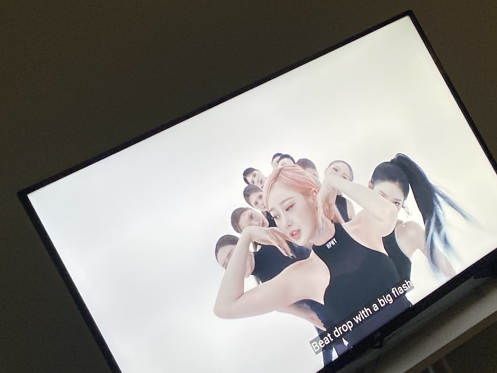 An image captures a television screen displaying a music video or performance. The screen is tilted slightly due to the angle of the photograph, revealing its black bezel, with a faint "JVC" logo visible on the bottom right. The television is set against a dark, featureless background, suggesting an indoor, possibly dimly lit, environment.
On the TV screen, several individuals, presumably female dancers, are arranged in a dynamic, staggered formation against a bright, plain white background. The most prominent figure, positioned towards the center-right, has light pink hair styled in an updo and wears a black, one-shoulder top with the text "HPNY" printed in white on the chest. She is captured in a pose with her arm raised, her expression focused or intense. Behind her, other dancers with dark hair, some styled in ponytails, are visible in various poses, also wearing black attire.
At the bottom of the screen, white subtitles are displayed against a dark, semi-transparent bar, reading: "Beat drop with a big flash". The overall scene on the television conveys a sense of high energy and performance.