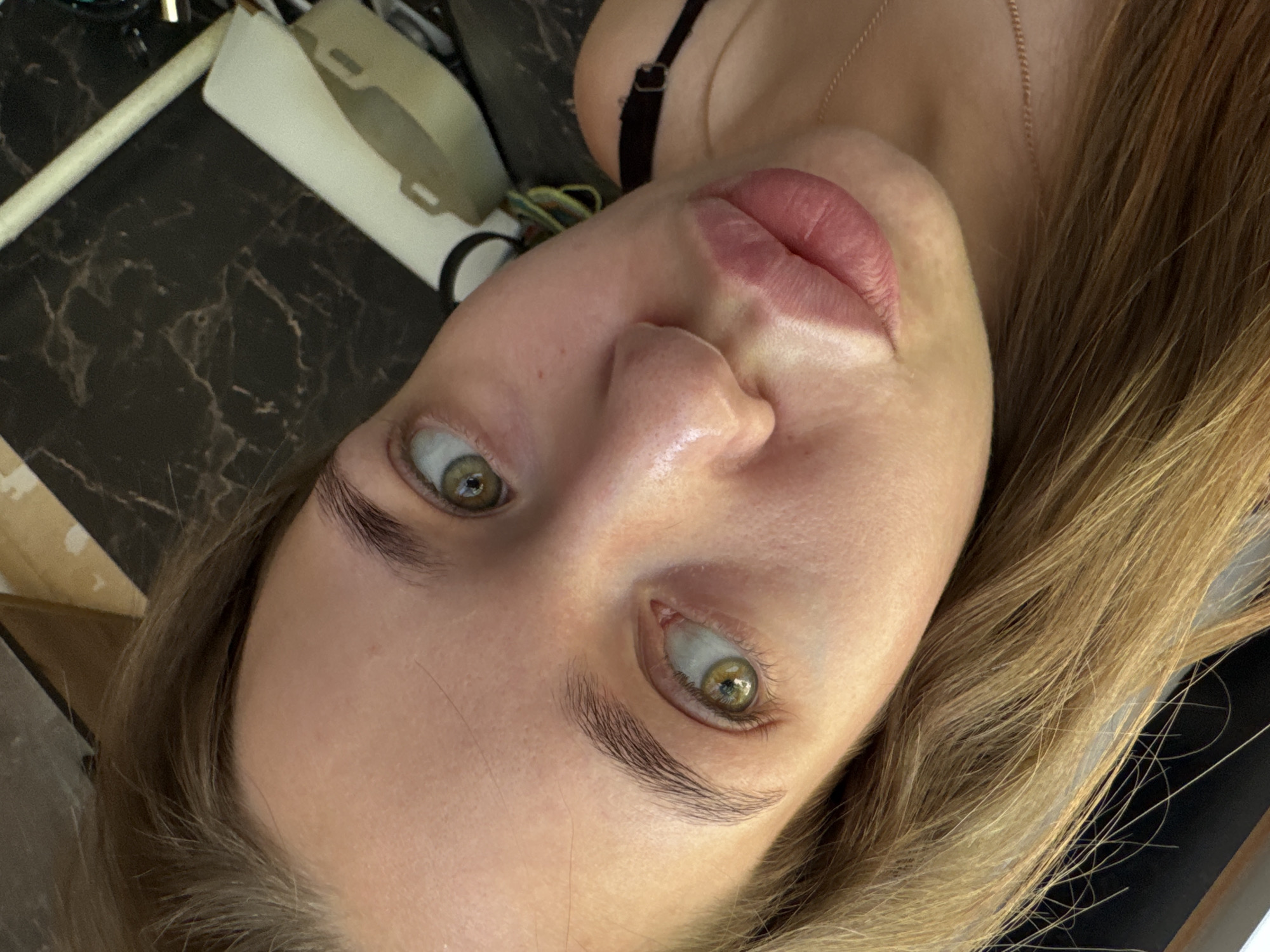 The image presents a close-up, angled portrait of a young woman looking directly at the camera. Her face is positioned diagonally, with her head tilted to the viewer's right. She has wide-open, striking green or hazel eyes and defined, light brown eyebrows. Her skin is fair, with a slight sheen visible on her forehead and the bridge of her nose. Her lips appear full, with a noticeable transition in color on her upper lip, suggesting a possible cosmetic treatment such as lip tinting or tattooing.
Her light brown or blonde hair frames her face, flowing down the right side of the frame. She is wearing a thin black strap, likely from a top or undergarment, and a delicate gold chain is visible around her neck.
The background is largely dark and appears to be an indoor setting. To the upper left, a dark surface with a faint white marble pattern is visible, upon which a light-colored cylindrical object and a light-colored, possibly foam or plastic, item are placed. The lighting is soft and appears to be directed towards her face, clearly illuminating her features. There is no discernible activity taking place other than posing for the photograph. Her expression is neutral, characterized by an intense direct gaze. No text is visible in the image. The location context provided is Chaikovsky, Russia.