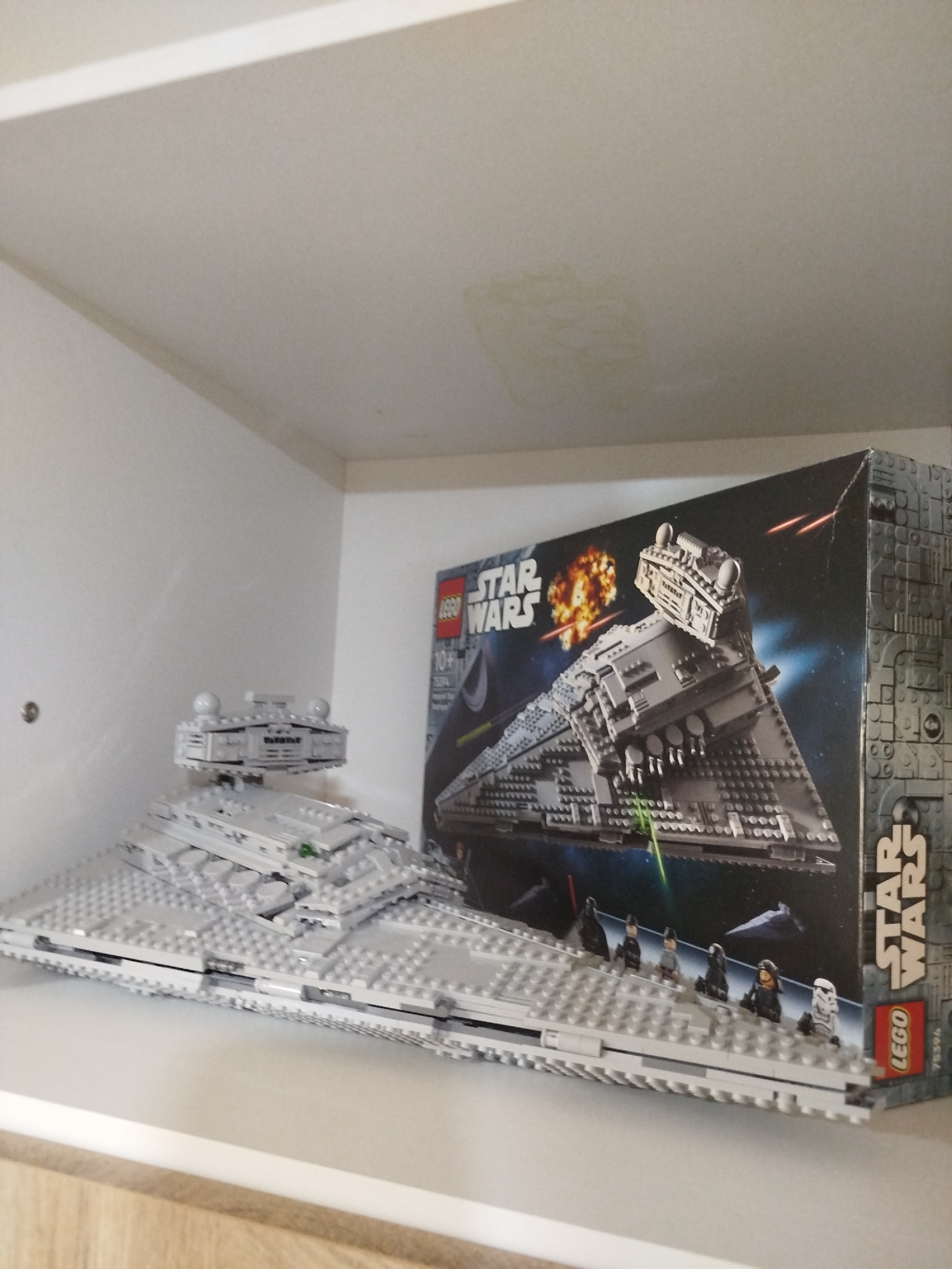 The image shows a Lego Star Wars Imperial Star Destroyer model and its box on a white shelf. The shelf is positioned within a room with white walls and a light wood-colored lower shelf visible at the bottom. The Lego model is predominantly gray and appears to be assembled. The box behind the model features an image of the Star Destroyer in space, with explosions and laser fire. The Lego logo and "Star Wars" branding are clearly visible on the box. There are no people present in the image. The lighting in the scene is diffused, suggesting an indoor environment. There is no information available to link this image to the city of Mirny, Russia.
