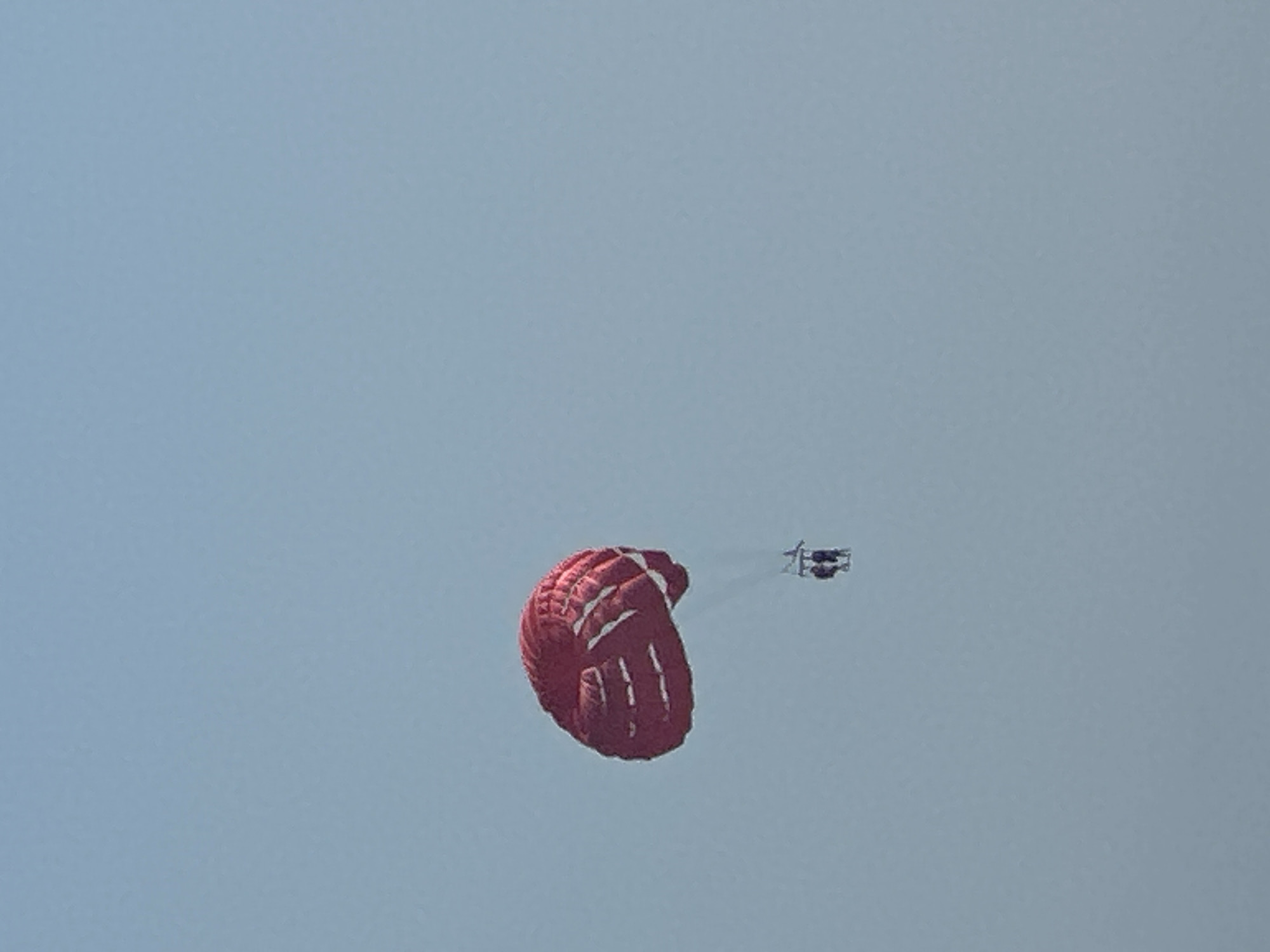 In Boreti, Montenegro, two individuals are paragliding in tandem, suspended from a large red parachute. The parachute, characterized by its bright red color and white stripes, is prominently featured against a clear, pale blue sky. The two people are seated in a harness suspended below the parachute, appearing to be enjoying the aerial activity. The overall scene depicts a recreational activity taking place in an open, outdoor environment, under a bright sky.