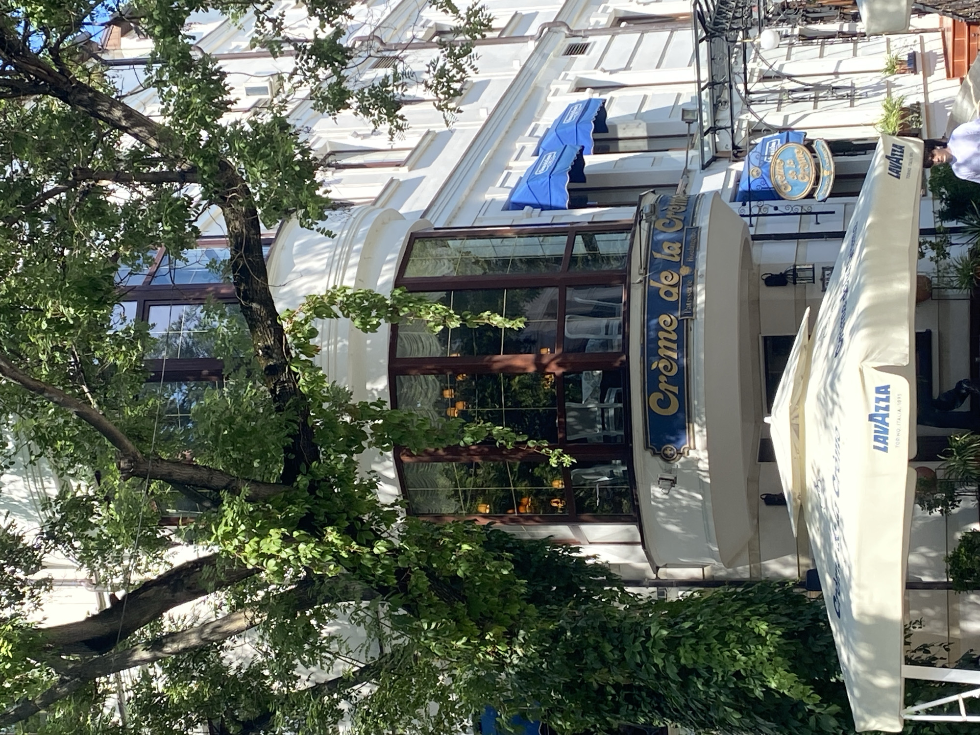 The image shows the exterior of a building in Chișinău, Moldova, featuring a cafe called "Crème de la Crème". A large tree with lush green foliage partially obscures the building's facade. On the first floor of the building, there are outdoor seating areas covered by umbrellas. One umbrella displays the "Lavazza" logo. The building itself is white with several windows, some of which have blue awnings. A sign with the cafe's name is prominently displayed. A mannequin is visible near the entrance, dressed in a white shirt. The lighting suggests it is daytime, with sunlight casting shadows.