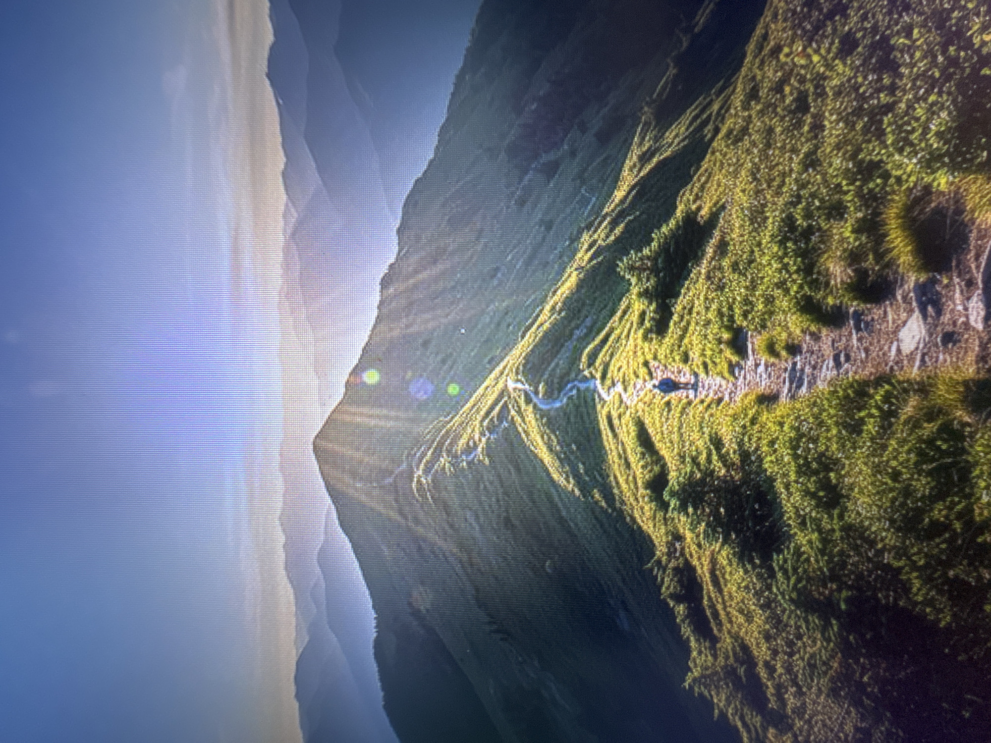 An individual walks along a winding path atop a grassy mountain ridge during sunrise. The sun's rays pierce through the sky, casting long shadows and illuminating the landscape. Layers of mountains recede into the distance, creating a sense of depth. The scene is located in the city of Stepnogorsk, Kazakhstan.