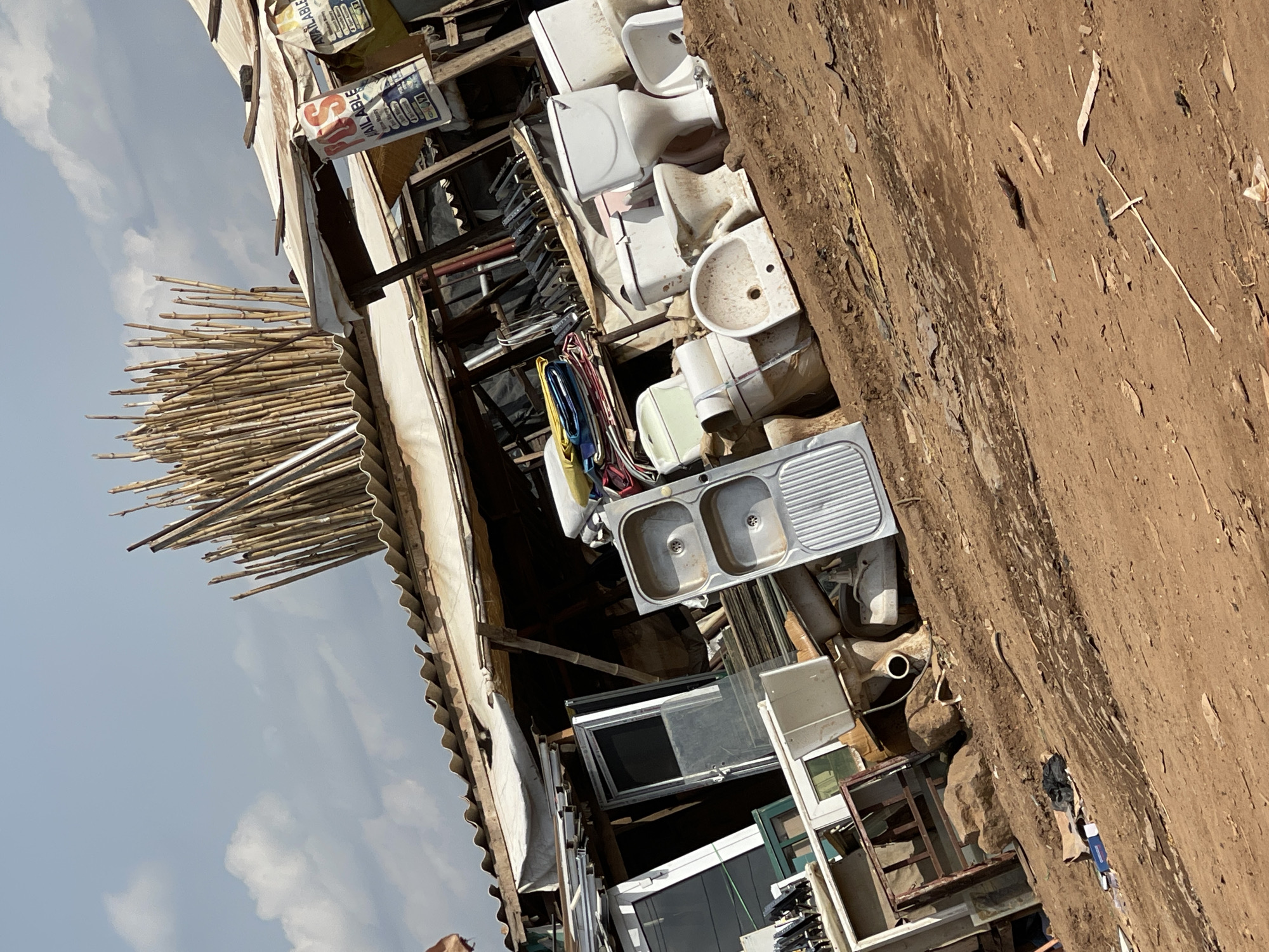 The image depicts a disorganized outdoor storage area in Abuja, Nigeria. The setting appears to be a construction site or a yard where discarded materials are kept.
Objects in the image include a double-basin stainless steel kitchen sink, multiple ceramic toilet bowls and cisterns, various window frames (some with glass panels), piles of bamboo poles, and building materials. Bags of cement or other construction products are stacked in the background.
The environment is characterized by exposed earth and what seems to be a partially constructed or dilapidated structure with a corrugated metal roof. The scene implies a temporary or transitional condition. The overall impression is one of salvaged, discarded, or stockpiled construction and household items.