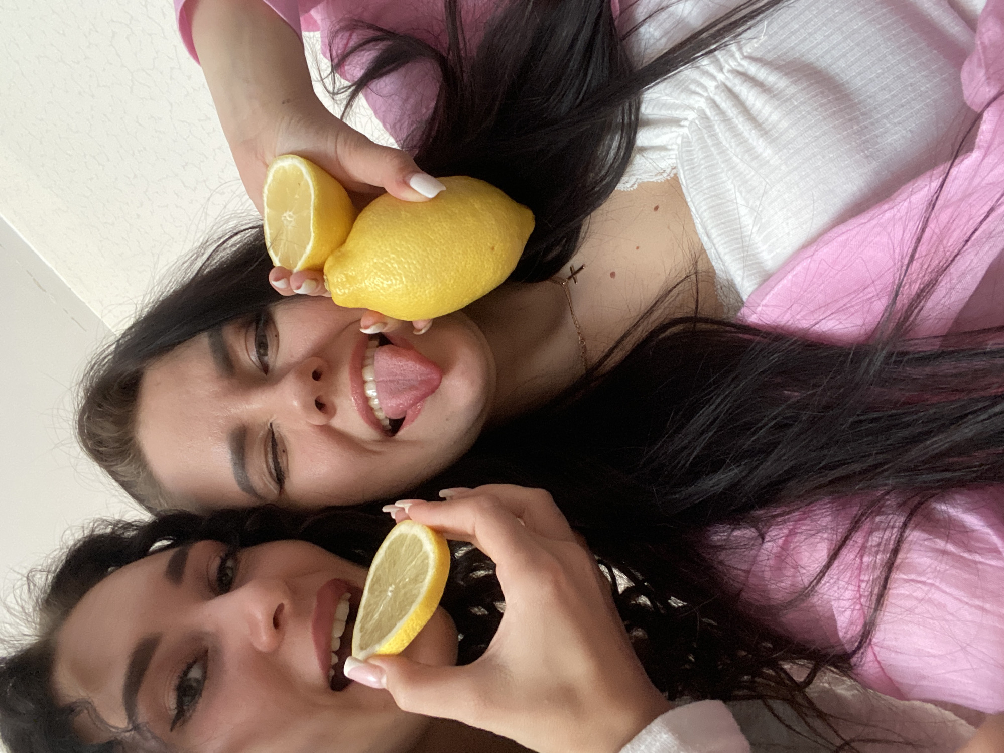 This close-up, upward-angled image captures two young women indoors, likely in Chaikovsky, Russia, engaged in a playful activity involving lemons. Both women have dark hair and are seen from a low perspective, as if the camera is looking up at them.
The woman on the right side of the frame holds a whole lemon and a half-lemon near her face. She has a wide, expressive smile, revealing her teeth, and her tongue is playfully sticking out. She appears to be wearing a light-colored, possibly white, ribbed top, and a subtle necklace with a small cross pendant is visible on her chest. Her nails are manicured with white tips.
The second woman, partially visible on the left side, also smiles broadly, showing her teeth, as she holds a half-lemon close to her mouth. Her dark hair frames her face, and her nails also feature a French manicure style. She is wearing a pink garment, which can be seen under her hair and as part of the background on the upper right.
The background is simple and light-colored, suggesting a plain wall or ceiling. The overall mood conveyed by the image is cheerful and lighthearted. No text is visible in the media file.