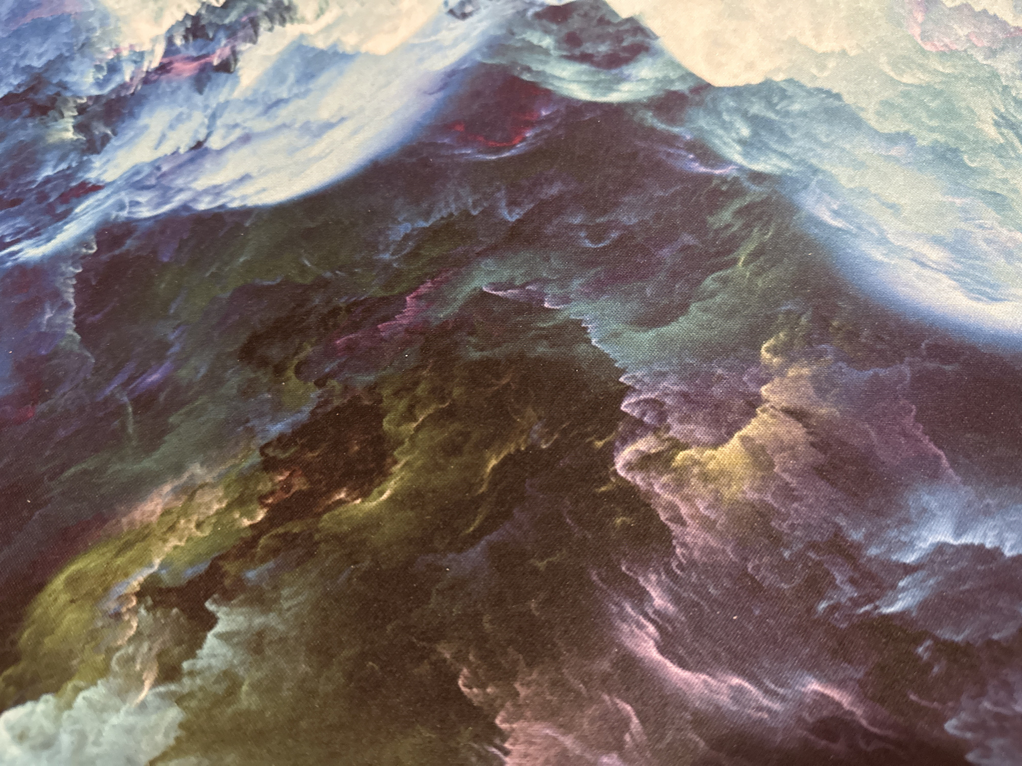 The image is a close-up, abstract shot of a fabric with a cloudy, nebula-like pattern. The colors are predominantly deep blues, purples, greens, and hints of yellow and pink, creating a swirling, ethereal effect. The texture of the fabric is visible, with a slight sheen and a weave pattern. The lighting is diffused, emphasizing the colors and shapes. There are no people or identifiable objects, and the location is unknown.