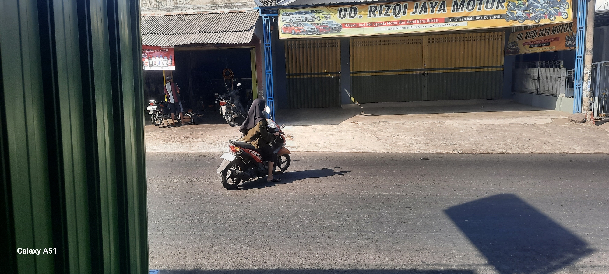 In Selopuro, Indonesia, a woman wearing a black hijab and a tan jacket rides a red and black scooter on a paved road. She is seen from the side, her face obscured by the hijab. The scooter is in motion, with its rear wheel visible. In the background, a shop called "UD. RIZQI JAYA MOTOR" is open, with a yellow corrugated metal facade. The shop appears to be a motorcycle dealership or repair shop, as several motorcycles are parked outside and inside. A man in a red and white shirt and blue shorts is standing near a parked motorcycle outside the shop. The environment is a street with a sidewalk in front of the shop. The sky is clear and the sun is casting shadows on the road. The overall scene depicts everyday life in a commercial area of Selopuro.