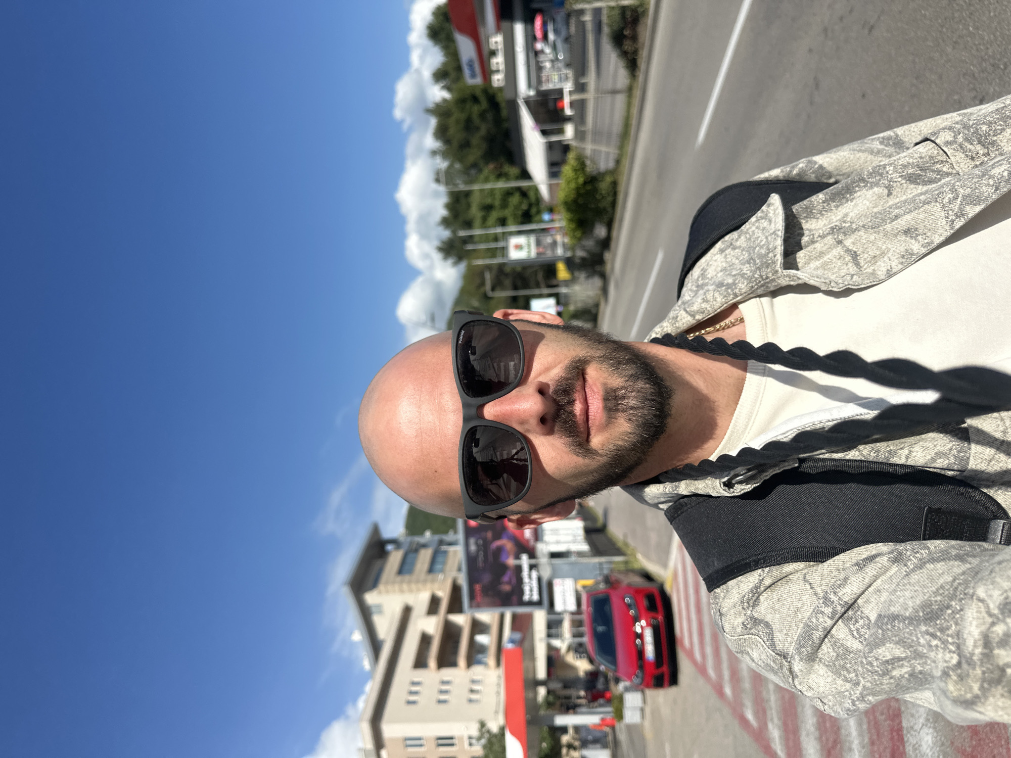 The image shows a close-up selfie of a bald man with a beard, wearing sunglasses, a white t-shirt, and a light gray jacket. The background shows a street scene in Budva, Montenegro, with a red car parked on a striped pedestrian crossing. In the background are buildings, a gas station, trees, and a clear blue sky with some clouds. The photo is taken during daylight hours.