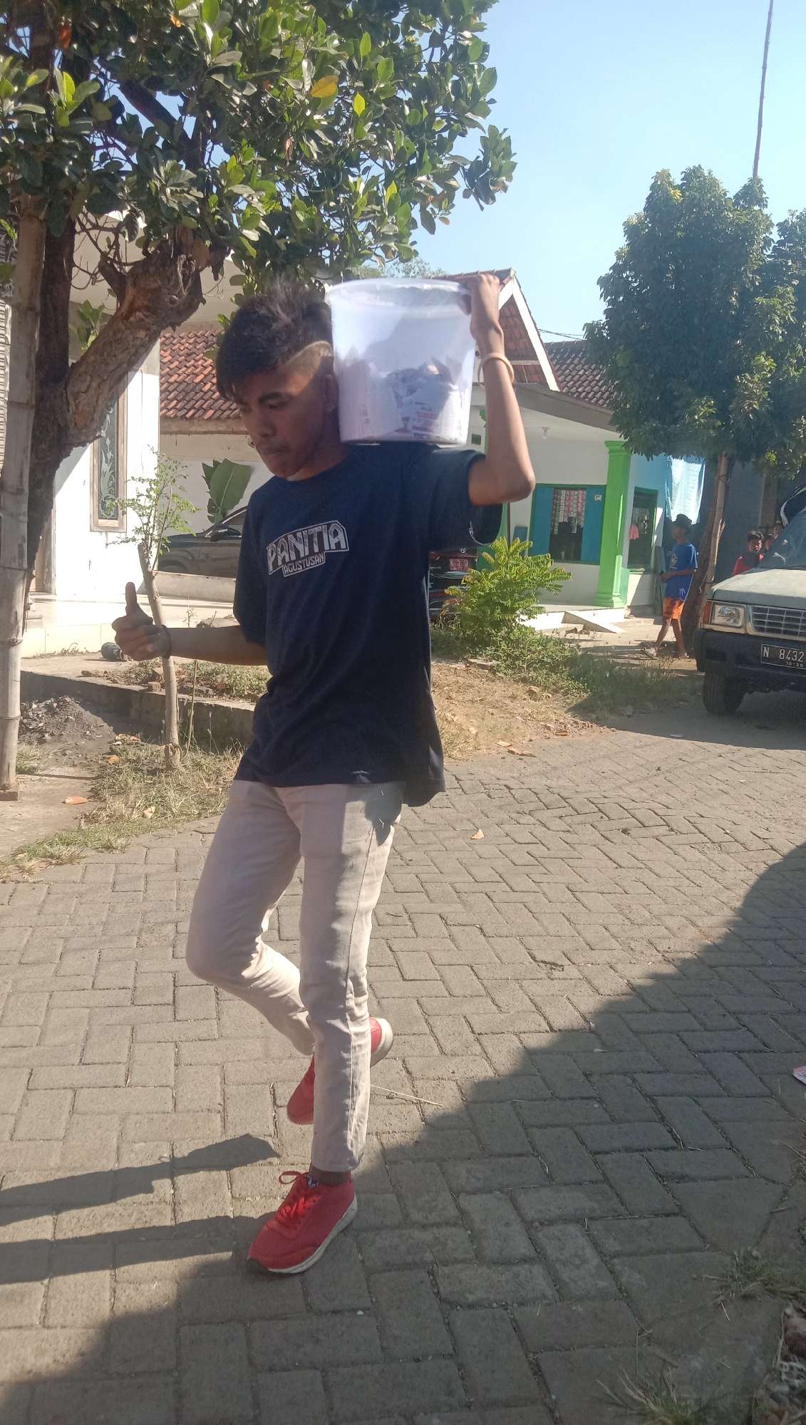 A young male, likely of Indonesian ethnicity, is pictured from the chest down, walking on a cobblestone path in Ngempit, Indonesia. He carries a clear plastic bucket filled with items on his right shoulder, supported by his right arm. His left arm is extended with his thumb up in a "thumbs up" gesture. He wears a dark blue t-shirt with white lettering that reads "PANITIA" and "AGUSTUSAN". His pants are light gray and he wears red athletic shoes. The background shows a residential area with houses, trees, and a parked car. The weather appears to be clear and sunny.