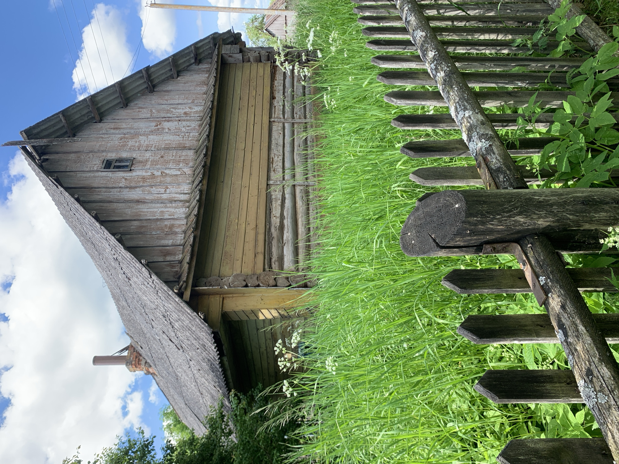 A weathered wooden house with a corrugated roof and a brick chimney with a metal extension stands amidst tall green grass and white flowering plants under a partly cloudy blue sky. The lower section of the house is constructed from horizontal logs, painted yellow-green, while the upper gable end is clad with vertical, unpainted, severely weathered wooden planks, featuring a small dark window. In the foreground, an old wooden picket fence, covered with lichen and featuring a rusted metal bracket, separates the viewer from the overgrown vegetation. To the far right, a utility pole with power lines is visible, along with a portion of another distant wooden structure. The scene is set in Відзаўскі сельскі Савет, Belarus, under bright daylight conditions.