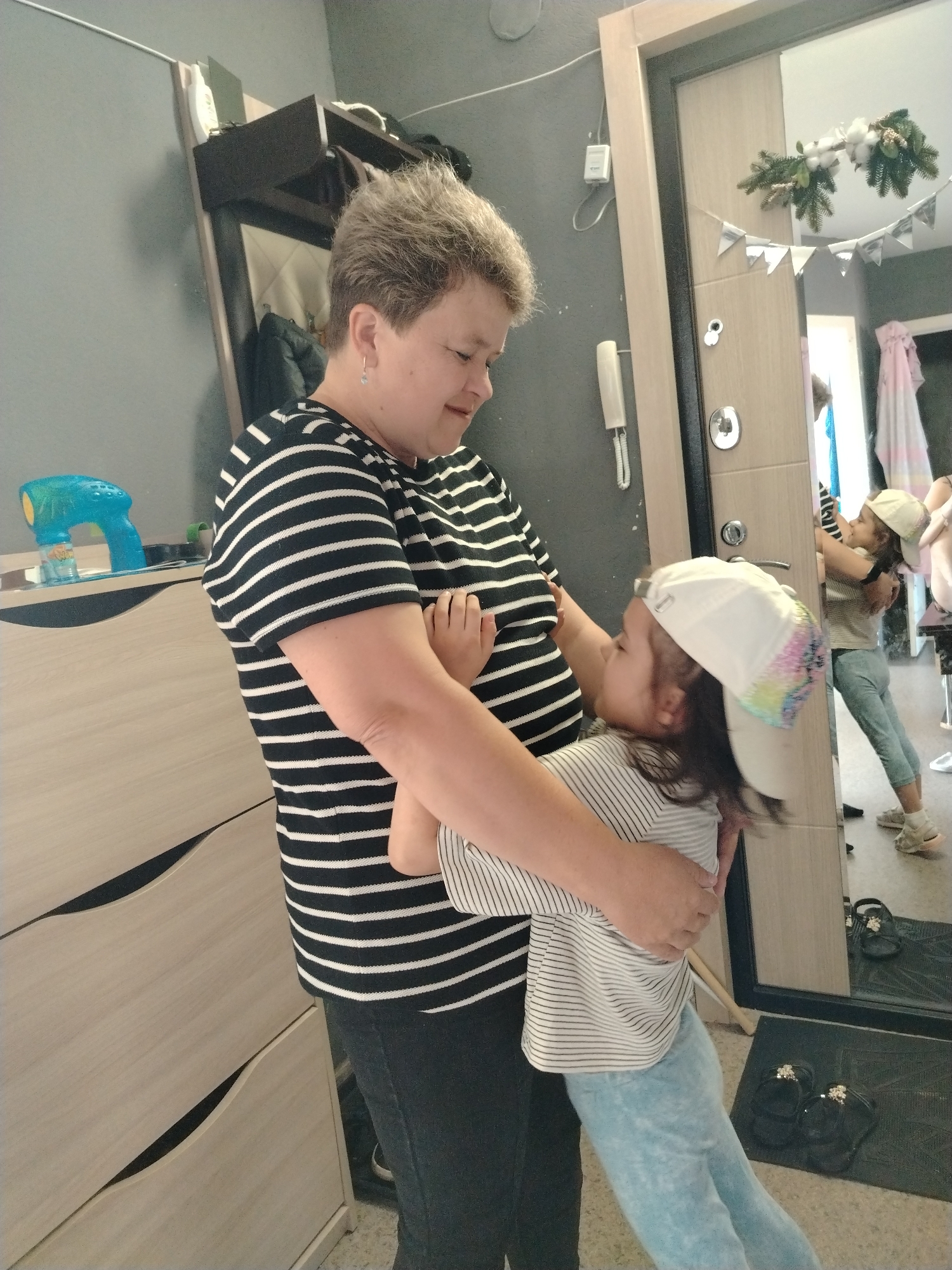 In Noviy, Russia, an adult woman with short, light brown hair and wearing a black and white striped t-shirt is holding a young child in her arms. The child is wearing a white baseball cap with colorful embellishments, a striped t-shirt, and light blue pants. The adult appears to be embracing the child, who is leaning into her. The scene takes place indoors, with a wall, a dresser with light-colored drawers, and a doorway with a mirror visible. In the mirror's reflection, another child, possibly the same one, is seen interacting with what appears to be a clothing rack. The environment suggests a residential or domestic setting.
