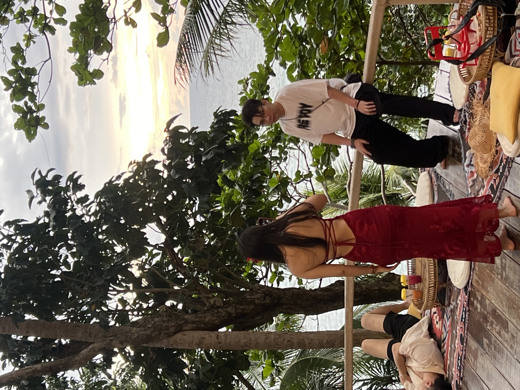 The image depicts three people on a wooden deck overlooking a body of water, possibly the ocean. The scene appears to be set in Karon, Thailand. A young man wearing a white t-shirt with the text "HEY JOY" and black pants stands looking towards the camera. Beside him, a woman in a long, red, backless dress is facing away from the camera, holding a phone. To the right, another person, possibly a man, is lying down on a rug, wearing a light-colored shirt and black shorts.
The time of day seems to be late afternoon or early evening, as indicated by the soft, warm light filtering through the trees and reflecting on the water. The sky is a hazy mix of yellow and white, suggesting a sunset or overcast conditions. Lush green foliage from trees and palm fronds frames the scene, creating a tropical atmosphere.
On the deck, several objects are visible, including a woven basket, a red chair, cushions, and a tray with drinks, suggesting a casual gathering or relaxation. The overall mood of the image appears to be relaxed and leisurely. The wooden deck and the natural surroundings point to an outdoor, possibly beachfront or elevated, location. The presence of multiple people on the deck suggests a social gathering or a shared moment of enjoyment in this scenic setting.