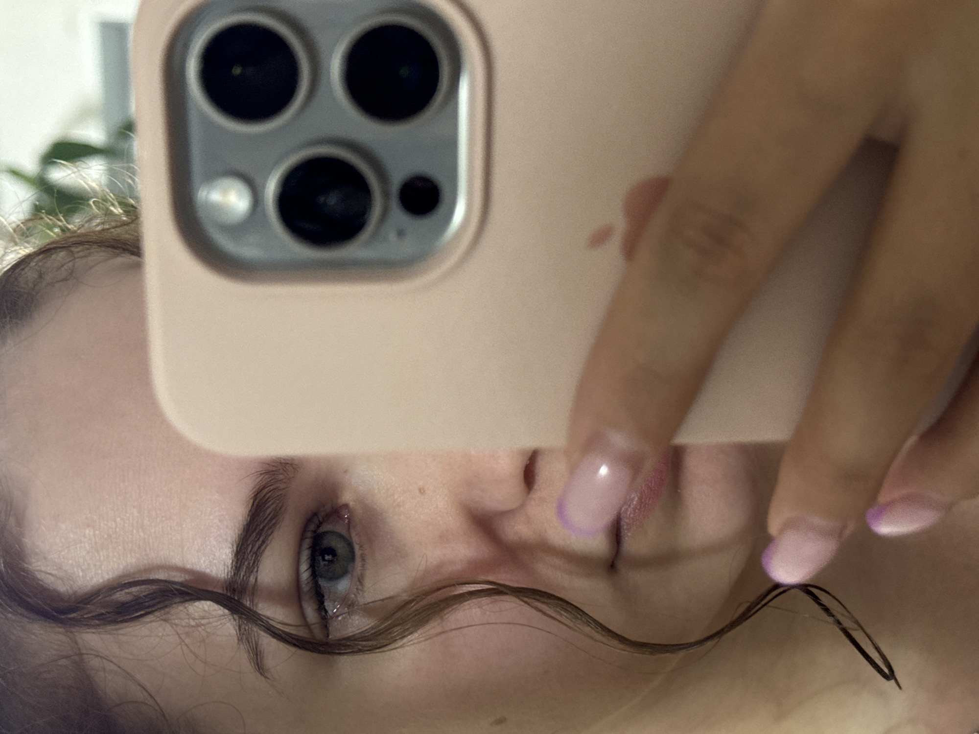 An indoor selfie captures a person's partially obscured face, taken from a slightly upward angle, with the image rotated 90 degrees counter-clockwise. The main subject is a person, likely female, whose left eye, eyebrow, and parts of their forehead, cheek, nose, and lips are visible. The eye has light-colored irises, possibly blue or grey-green, and shows a slight redness or irritation near the tear duct. Dark, wavy strands of hair frame the face.
A hand, possibly the subject's, holds a smartphone in a pale pink or peach-colored case, covering the upper part of the face. The phone case features a prominent camera module with three large lenses and a smaller flash component, indicative of an iPhone. A faint imprint of the Apple logo is visible on the case. The visible fingers of the hand have manicured nails, styled with a French manicure featuring light pink bases and purple tips.
The background is softly blurred, showing hints of what appears to be a houseplant and a bright, indistinct area, suggesting an indoor setting with diffused light. The activity depicted is the act of taking a self-portrait. There is no explicit text visible other than the subtle Apple logo on the phone case. The scene is located in Chaikovsky, Russia.