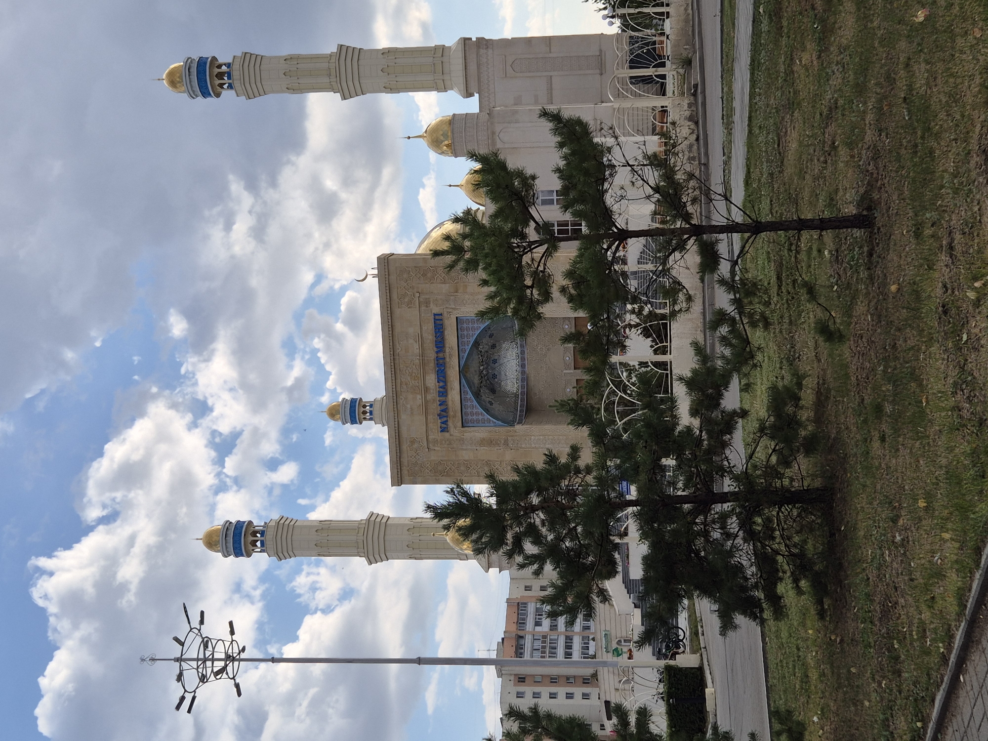 The image shows the Nur-Mubarak Mosque in Kokshetau, Kazakhstan. The mosque is a large, modern building with a light-colored stone facade. It features two tall minarets topped with golden domes and blue accents, as well as several smaller golden domes. The main entrance is an arched opening with intricate carvings above it. The text "NATAN HAZIRET MESCITI" is visible above the entrance. In front of the mosque, there are two small pine trees and a white decorative fence. To the left of the mosque, a tall street light with multiple lamp fixtures is visible. In the background, a multi-story apartment building can be seen. The sky is partly cloudy, with white clouds against a blue sky. The ground in front of the mosque is covered with grass and some fallen leaves.