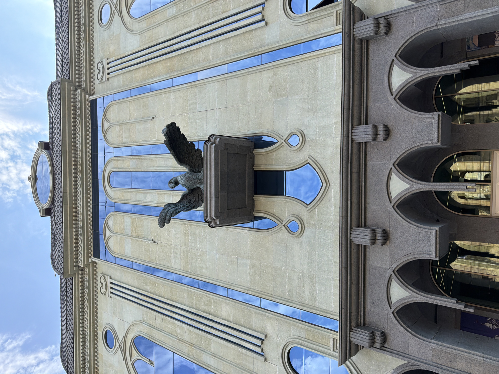 A low-angle shot captures the facade of a building in Tbilisi, Georgia, adorned with architectural elements reminiscent of Neo-Gothic and Art Nouveau styles. The building is primarily constructed of light-colored stone, with accents of darker stone on the lower levels. A prominent feature is a bronze eagle statue with outstretched wings, perched on a decorative ledge above a series of tall, arched windows. These windows are framed by a darker material and feature blue-tinted glass that reflects the sky. Vertical decorative elements resembling fluted columns run up the facade, interspersed with arched windows and circular window details. The roofline is detailed with cornices and decorative molding, topped by a dark, tiled roof and an oval dormer window. Below the main facade, a row of arched openings with dark stone framing and ribbed detailing suggests a covered walkway or entrance area. The sky above is a clear blue with scattered white clouds.