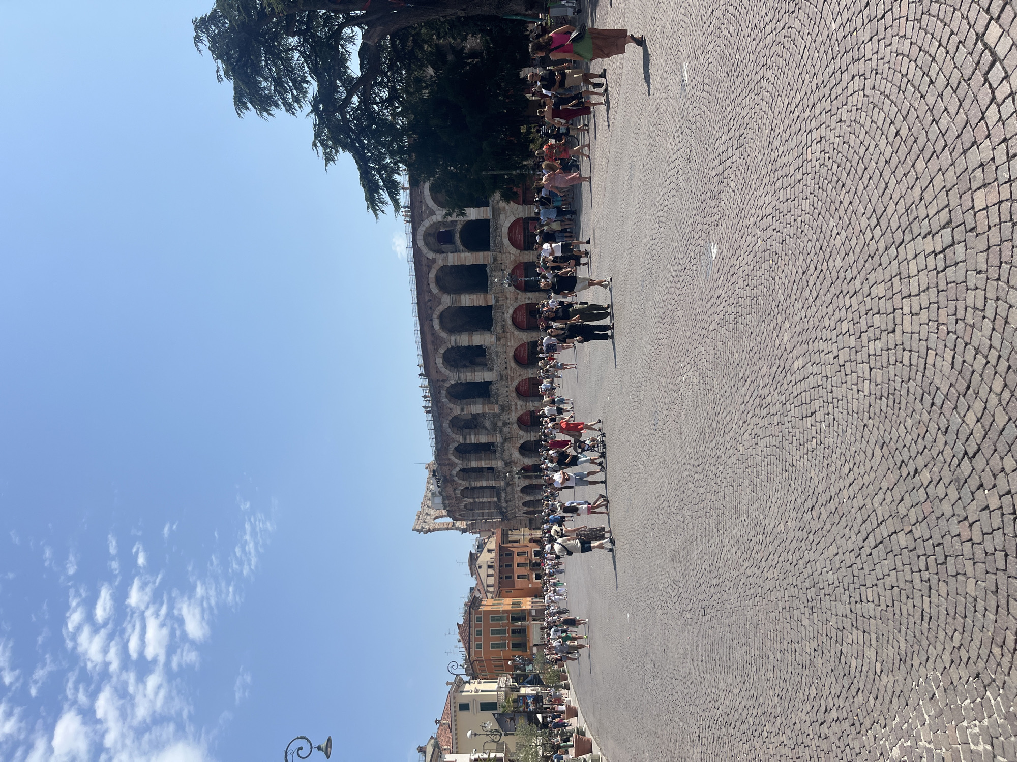 The image depicts a large, open, cobblestone plaza in front of an ancient Roman amphitheater, the Arena di Verona, in Verona, Italy. The sky is a clear, bright blue with scattered white clouds. The amphitheater, a prominent architectural structure with arched openings and weathered stone, dominates the right side of the frame. Numerous people are scattered across the plaza, walking, standing, and interacting. They are dressed in casual summer clothing.
Buildings with orange and cream-colored facades line the left side of the plaza, suggesting a vibrant urban setting. Several lampposts with decorative tops are visible. The cobblestone pavement is laid in a circular pattern, creating a visually dynamic foreground. The overall scene is one of a bustling tourist destination on a sunny day, with the historical Arena di Verona serving as the central focal point. A large tree with dense foliage stands to the right of the amphitheater, partially obscuring it.