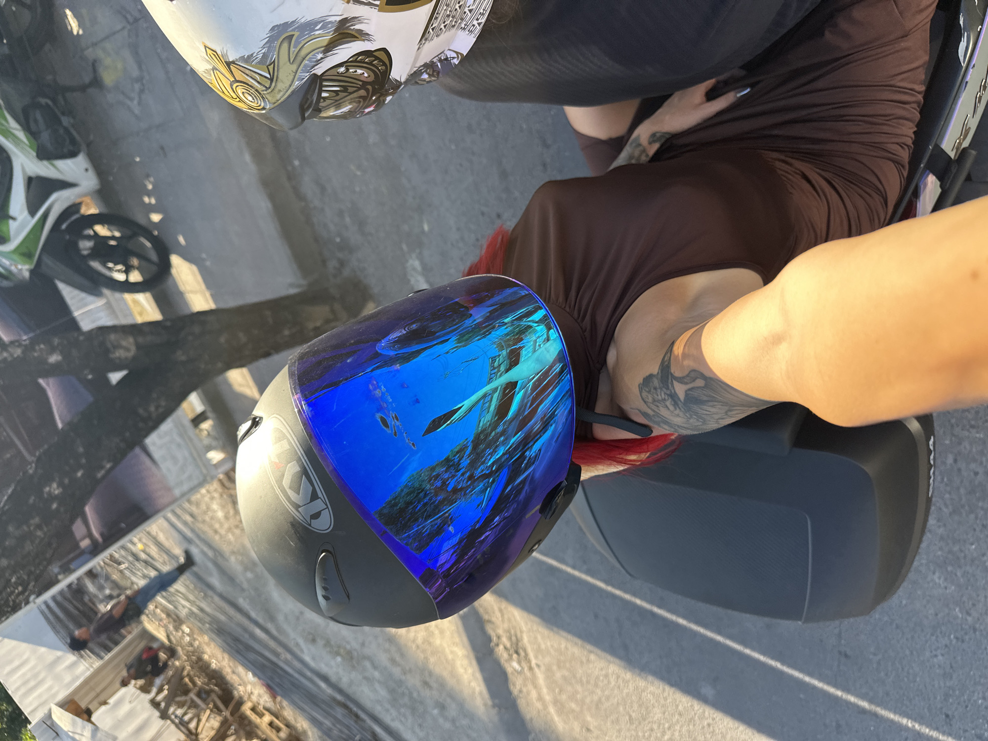 A close-up, angled shot depicts two people on a motorbike in North Kuta, Indonesia. The person on the back wears a brown dress and a black helmet with a blue mirrored visor. The other person has a white helmet. The background shows a paved surface with shadows.