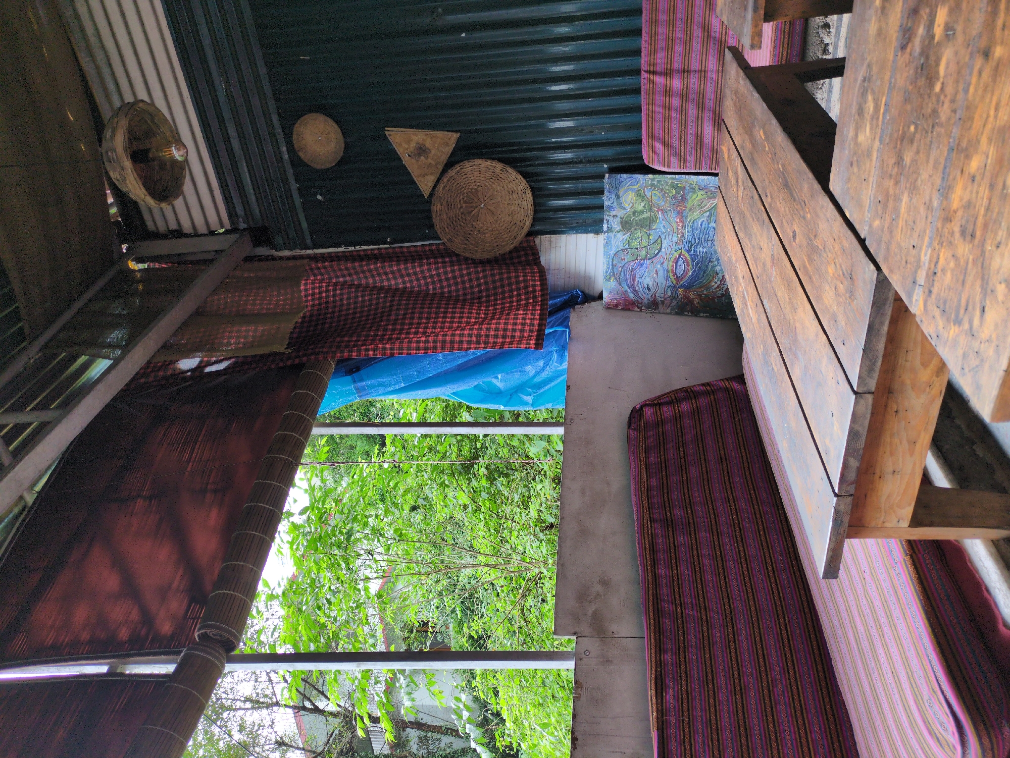 The image depicts an outdoor seating area, possibly a cafe, with rustic wooden tables and benches covered in striped purple and pink fabric. A large window overlooks lush green trees and foliage. The interior wall is made of corrugated metal, decorated with woven circular objects and a triangular piece of wood. A woven pendant light hangs from the ceiling. A blue tarp and red plaid fabric are draped over a beam, adding to the informal atmosphere. A colorful abstract painting is propped against the wall. The overall impression is one of a casual, bohemian space. The scene is located in Bhagsu, India.