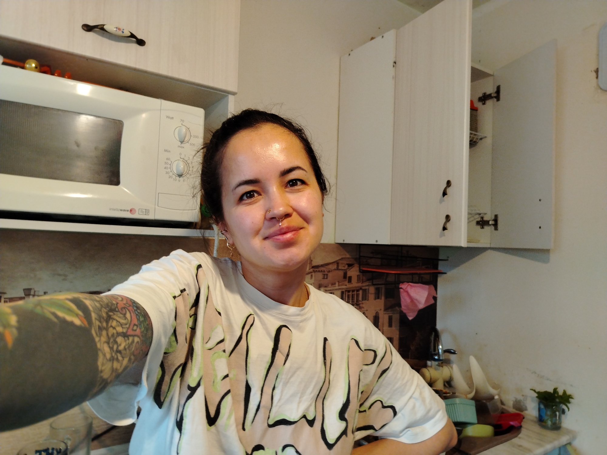 A woman is in a kitchen. She is fair-skinned with dark hair pulled back and a nose ring. She wears a white t-shirt with black and beige designs. Her left arm is heavily tattooed. Behind her is a white microwave and white cabinets, one of which is open. A picture hangs behind the counter, along with various kitchen items. There is a plant on the counter to the right.
The image does not provide information about a location, including the city of Noviy, Russia.