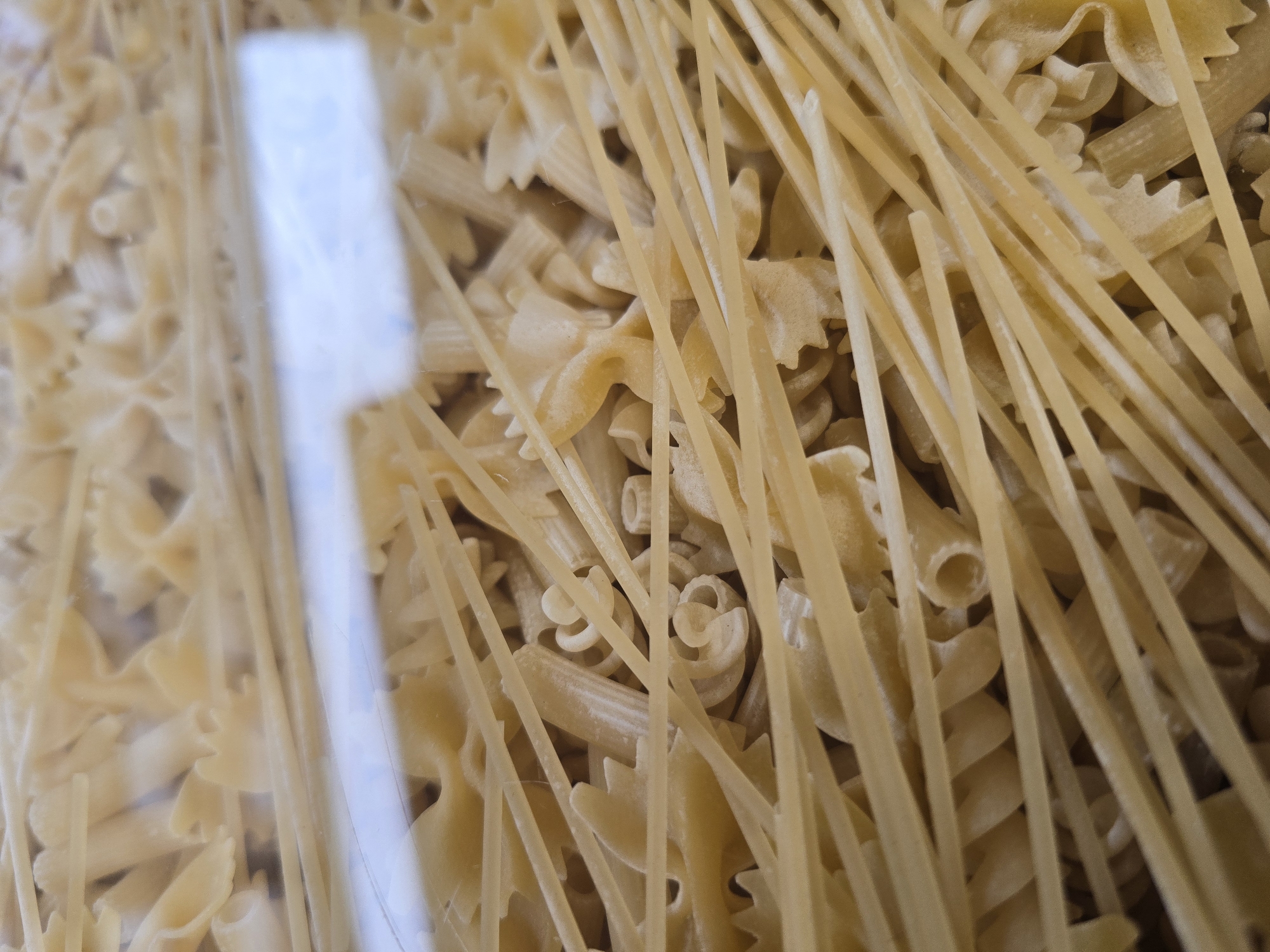 The image displays a close-up, overhead view of various types of dry, uncooked pasta. The dominant feature consists of numerous long, thin, light-yellow strands resembling spaghetti or linguine, arranged diagonally across the frame. Beneath and interspersed among these strands are different pasta shapes, including farfalle (bow-ties), fusilli (spirals), and tubular pasta pieces consistent with penne or rigatoni. A fine dusting of white powder, likely flour or pasta residue, is visible on many surfaces. The pasta is contained behind a clear, reflective barrier, possibly plastic or glass, which exhibits a horizontal band of blurred reflection in the central mid-ground. No individuals, actions, or specific environmental details beyond the pasta container are visible. The precise location within the city of Sofia, Bulgaria, cannot be determined from the visual information provided.
