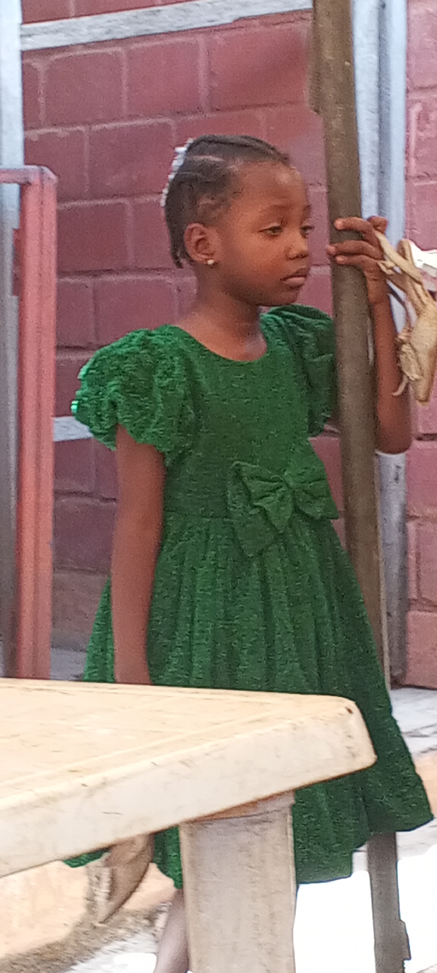A young girl wearing a green dress with a bow stands next to a pole. The location is an urban setting, possibly in the city of Unknown, Unknown, indicated by the brick wall in the background and the edge of a table in the foreground. The girl has braided hair and is wearing earrings. She is holding onto the pole with her left hand. The environment appears to be outdoors.