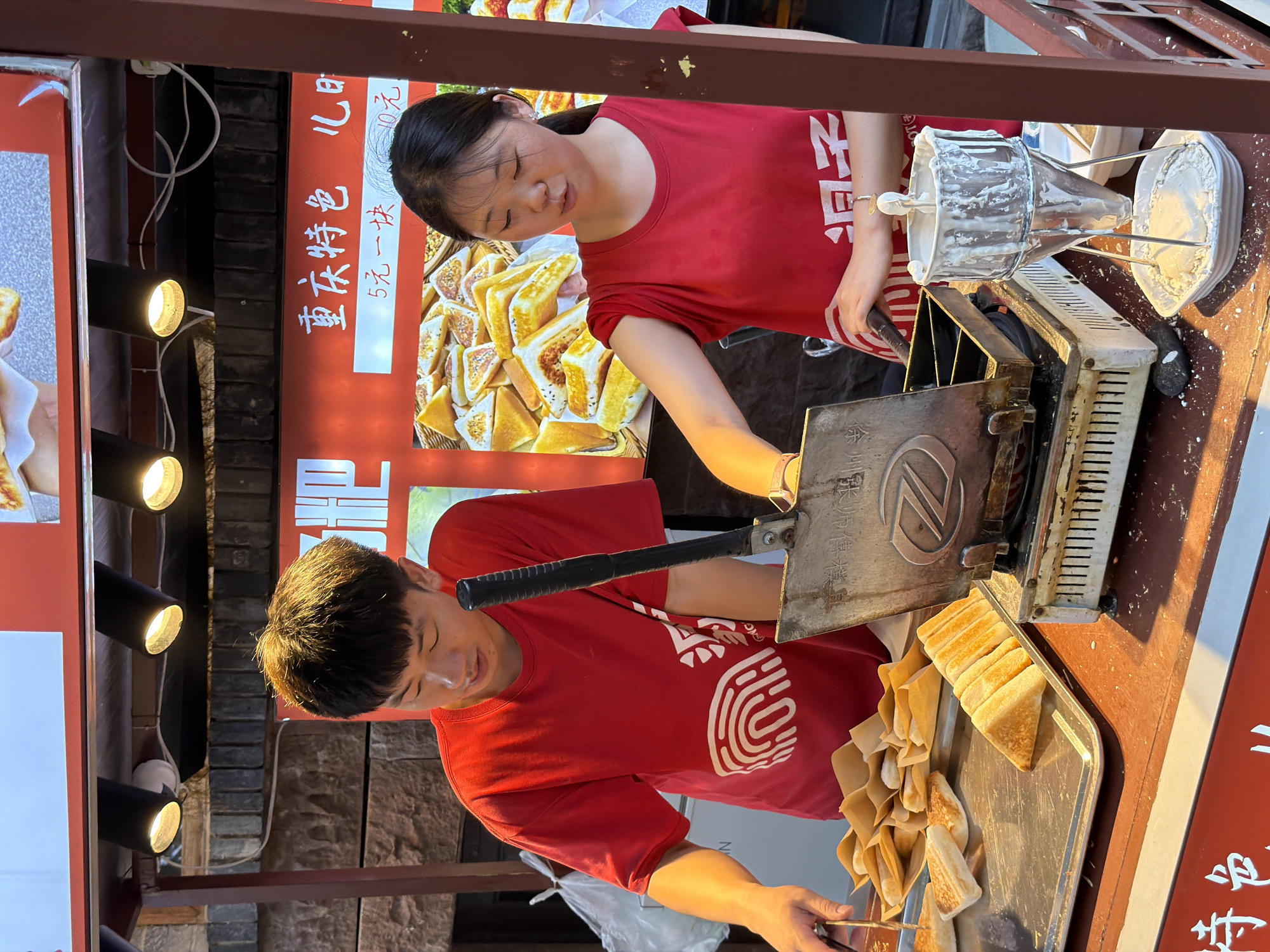 Two young people, a man and a woman, are working at a food stall in Yuzhong District, China. They are both wearing red t-shirts. The woman, standing behind a food warmer, is leaning over a waffle maker. The man, in front, is preparing to place something onto a baking tray. A sign behind them displays text and images of toasted sandwiches, indicating they are selling a type of street food. The sign also has prices written on it. The scene appears to be outdoors, possibly in a busy market or on a street, as suggested by the lighting and the materials of the stall. The overall atmosphere seems to be one of active commerce, with the two individuals focused on their tasks. The lighting suggests it could be daytime or early evening.