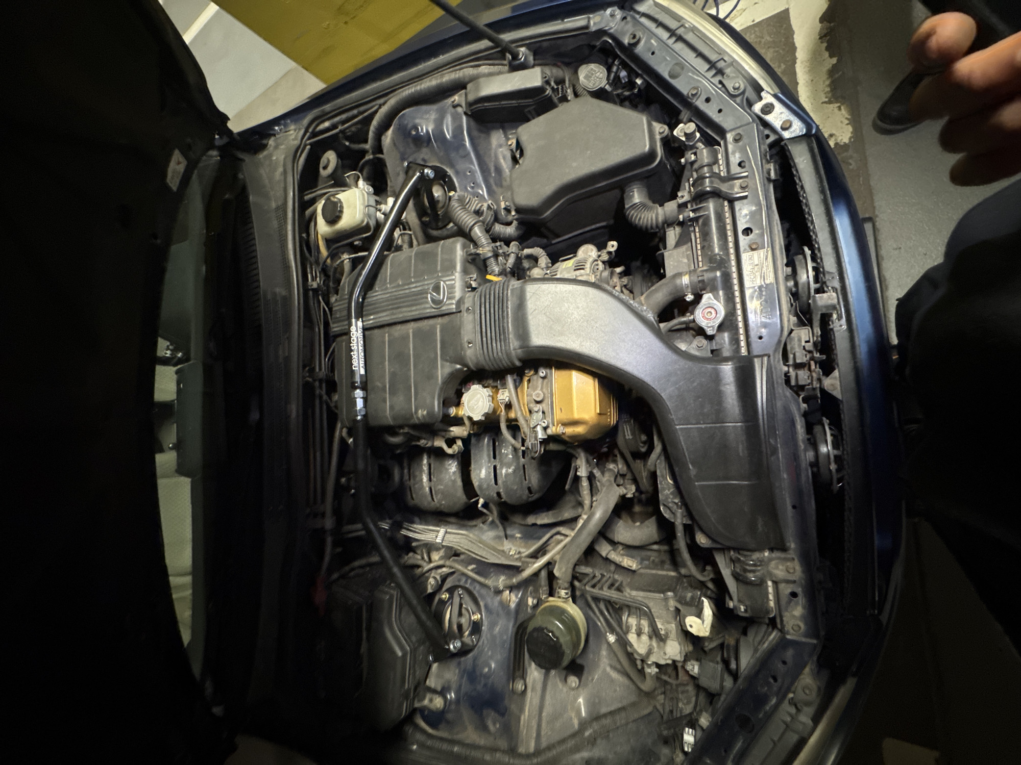 The image provides a detailed view of an opened car engine bay, belonging to a blue Lexus vehicle, with the hood partially visible on the left. The engine compartment is densely packed with various components. A black engine cover prominently displays the "Lexus" logo. Above it, a brake fluid reservoir is visible. A black aftermarket strut bar, adorned with the white text "next stage Automotive," spans across the upper engine bay, indicating modifications or performance enhancements.
Towards the front right, a radiator and a red-capped coolant reservoir are present. Numerous hoses, wires, and black plastic components, including a substantial air intake system, are interconnected throughout the bay. A distinctive gold-colored component, possibly an aftermarket intake manifold, stands out towards the lower right of the main engine cover. The text "CALSONIC KANSEI" is partially visible on a cap of this gold-colored part. A warning triangle with an exclamation mark is on the coolant cap.
The setting appears to be an indoor space, likely a garage or workshop, given the artificial lighting and the visible concrete or tiled floor in the background. A human hand is visible on the far right, suggesting a person is interacting with or observing the engine. The engine bay exhibits some signs of use, including a light accumulation of dust and grime. This scene is located in Sofia, Bulgaria.