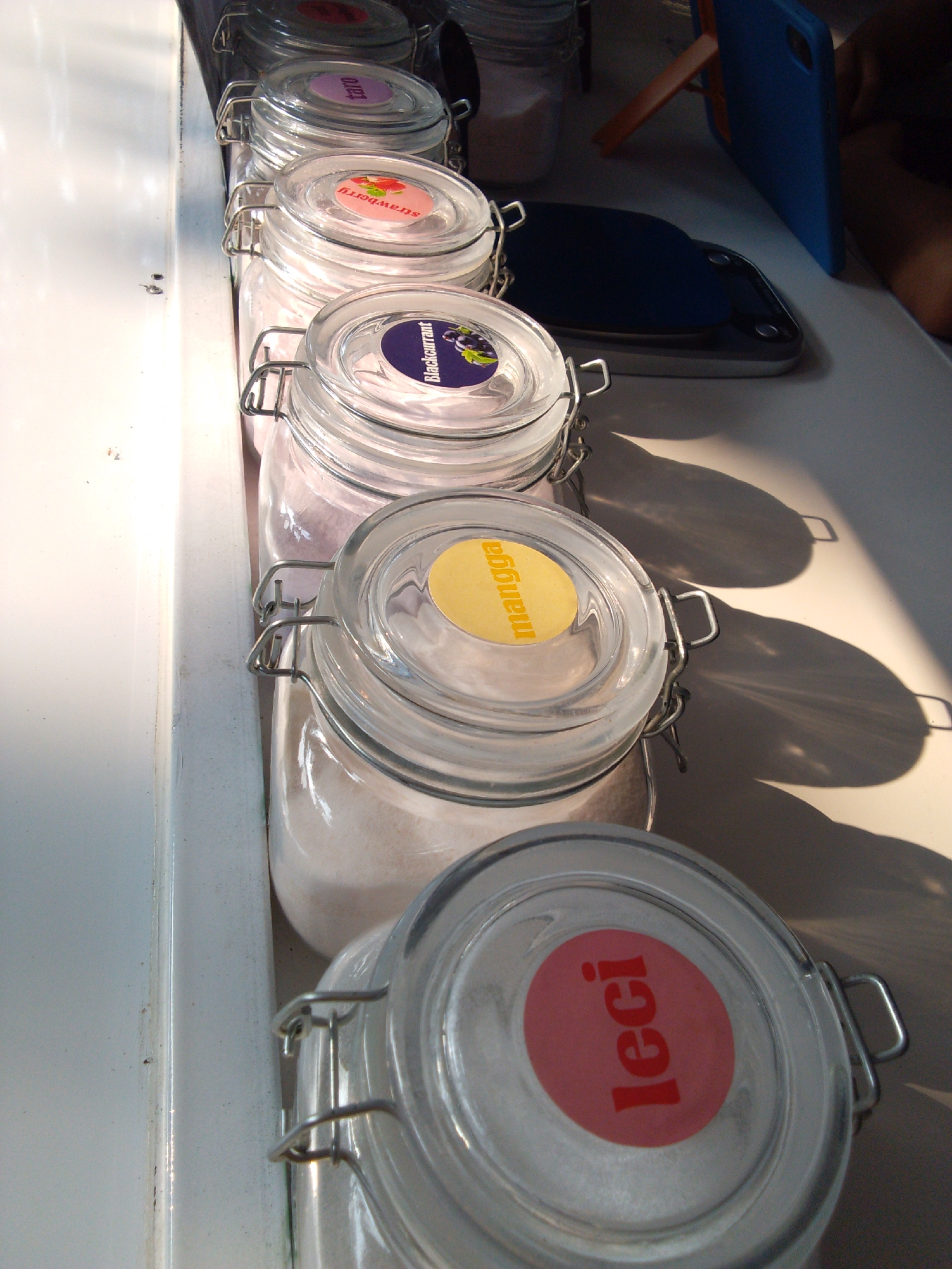 The image shows a row of glass jars with metal clasps, lined up on a white surface. Each jar has a lid with a circular label. From top to bottom, the visible labels read: "strawberry," "blackcurrant," "mangga," and "leci." The jars appear to contain a light-colored powder or granular substance. In the background, a blue tablet is propped up on an orange stand. Sunlight is casting distinct shadows on the white surface, suggesting an outdoor or well-lit indoor setting. The specific location in Trenggalek, Indonesia cannot be determined from the image alone.