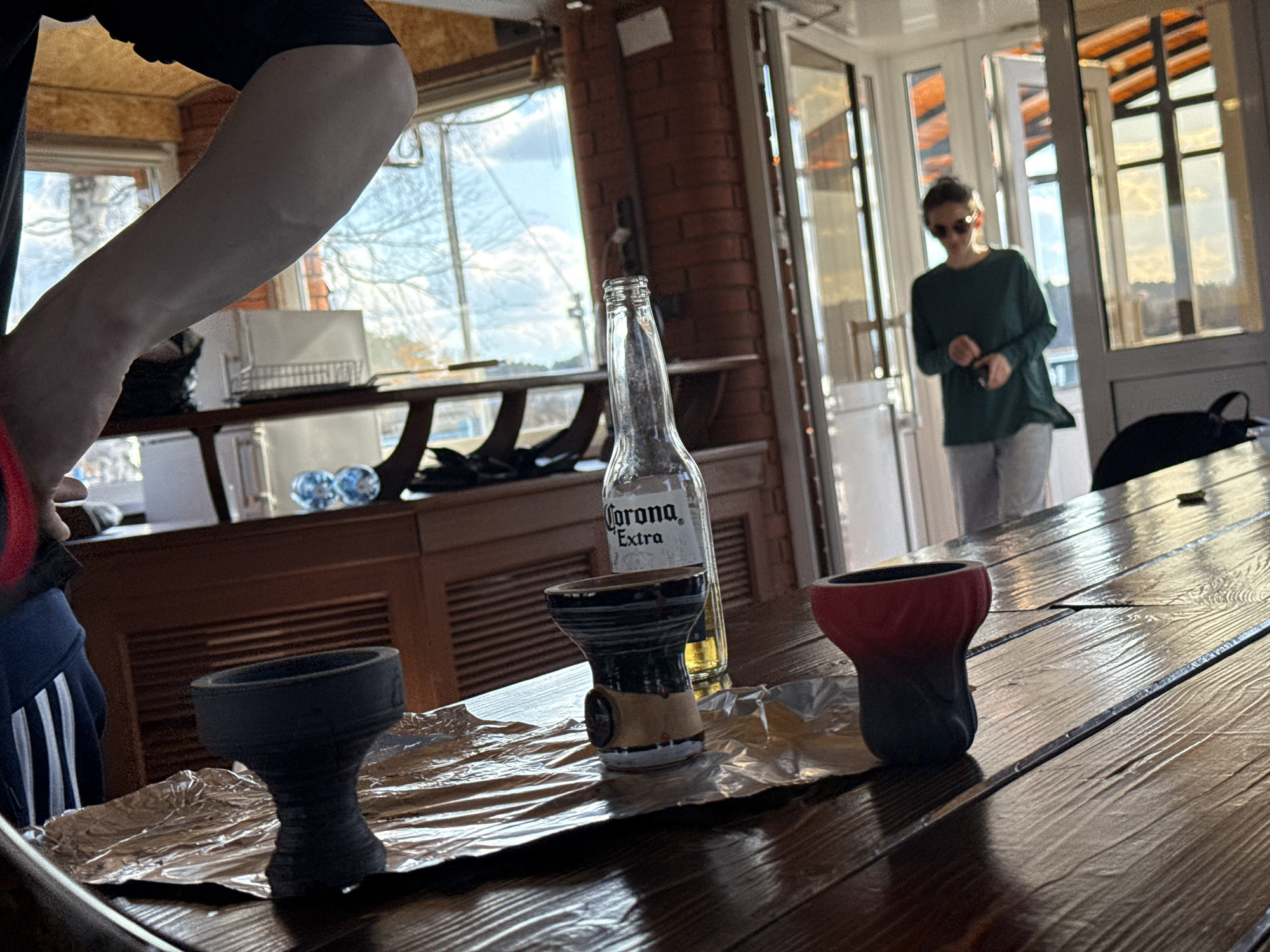This image depicts an indoor scene, possibly a lounge, cafe, or a large communal room, characterized by natural light and a mix of rustic and modern elements. In the foreground, a dark, glossy wooden table is prominent, upon which three ceramic hookah bowls of varying designs (one black, one patterned black/tan, and one red/black gradient) are placed on crinkled aluminum foil. A clear glass bottle of "Corona Extra" beer stands behind the patterned bowl.
To the left, part of a person is visible, including an arm, torso in a dark top, and blue track pants with white stripes. Their face and specific activity are not discernible. In the background, a blurred figure, wearing sunglasses, a dark green long-sleeved top, and light pants, walks towards the viewer from a brightly lit area featuring large glass doors and windows. This area allows ample natural light into the room, suggesting it is daytime with a clear sky visible outside.
The setting includes exposed brick pillars and wooden ceiling beams, creating a warm and inviting atmosphere. Behind the main table, a dark wooden cabinet holds a white appliance, likely a microwave, and a white dish drying rack, along with some decorative items. The presence of hookah bowls and the beer bottle suggests a casual social gathering or the preparation for one. The visible text on the bottle reads "Corona Extra." The location context of Chaikovsky, Russia, aligns with the style of a modern yet comfortable establishment, though no explicit visual cues within the image directly denote this specific location.