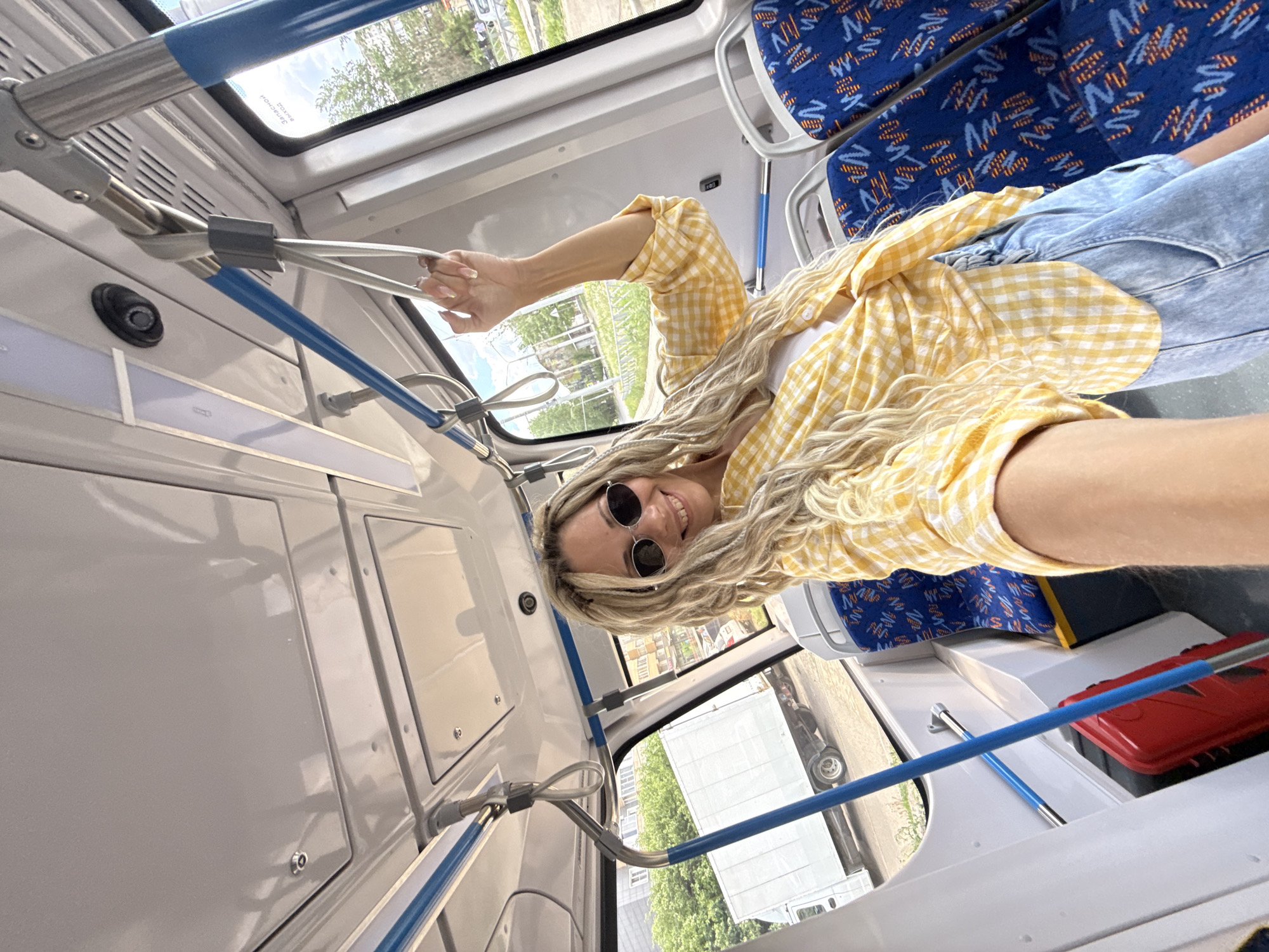 A blonde-haired woman wearing a yellow checkered shirt, light blue denim shorts, and round sunglasses takes a selfie on a bus. She is holding onto a silver support bar. Blue patterned seats and a red object can be seen in the background. Outside the bus window, trees and buildings are visible. The image is taken inside a bus in the city of Rusanovo, Russia.