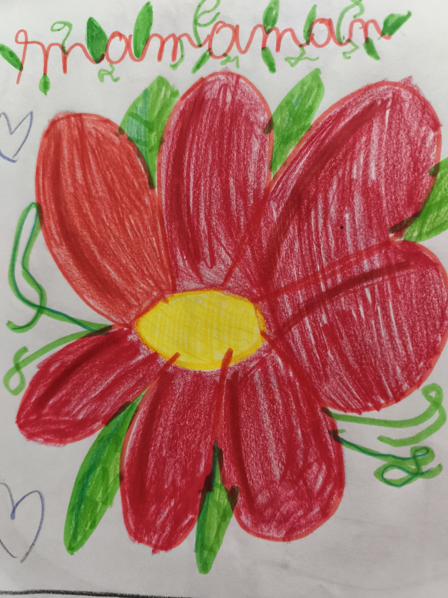 The image is a crayon drawing on white paper. The drawing depicts a large red flower with a yellow center. There are green leaves and vines surrounding the flower, as well as a few blue hearts drawn on the paper. At the top of the drawing, there is some red cursive writing, partially obscured by green drawings, which appears to read "maman." The drawing is amateurish and likely created by a child. The image itself does not provide any information about its location in Saint-Denis, France.
