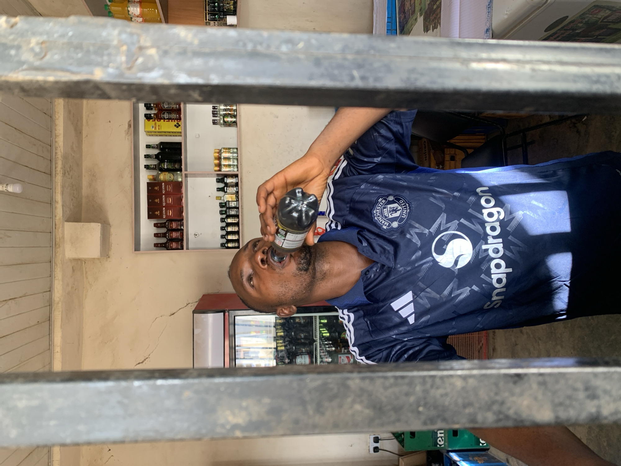 A man is drinking from a bottle, apparently inside a store or shop in Kabong, Nigeria. The man is Black, with a beard, and is wearing a blue soccer jersey with "Snapdragon" printed on it. He is holding a dark bottle to his mouth. Behind him is a refrigerated display case filled with beverages, and shelves stocked with bottles, likely alcoholic beverages. The view is partially obstructed by a metal security gate or bars. The floor appears to be concrete.