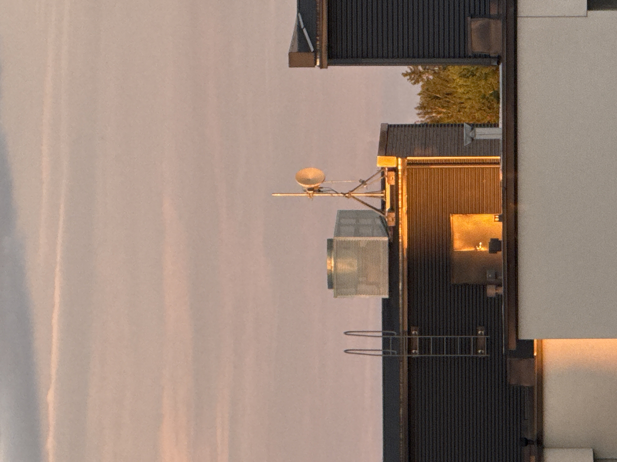 The image shows a rooftop scene at sunset. In the foreground, a white building with a dark railing is visible. Behind it, a darker building with a corrugated facade extends across the frame. On top of this building, a metal ladder leans against the facade. To the right of the ladder, a large, rectangular, metallic structure with vents is situated. Further to the right, on another section of the roof, a communication antenna with a satellite dish is mounted on a tripod. The antenna and dish are illuminated by the setting sun. In the background, trees with green and yellow foliage are visible, and the sky is filled with soft, pink and gray clouds, indicative of a sunset. The overall impression is one of an urban or industrial rooftop setting bathed in the warm light of dusk.