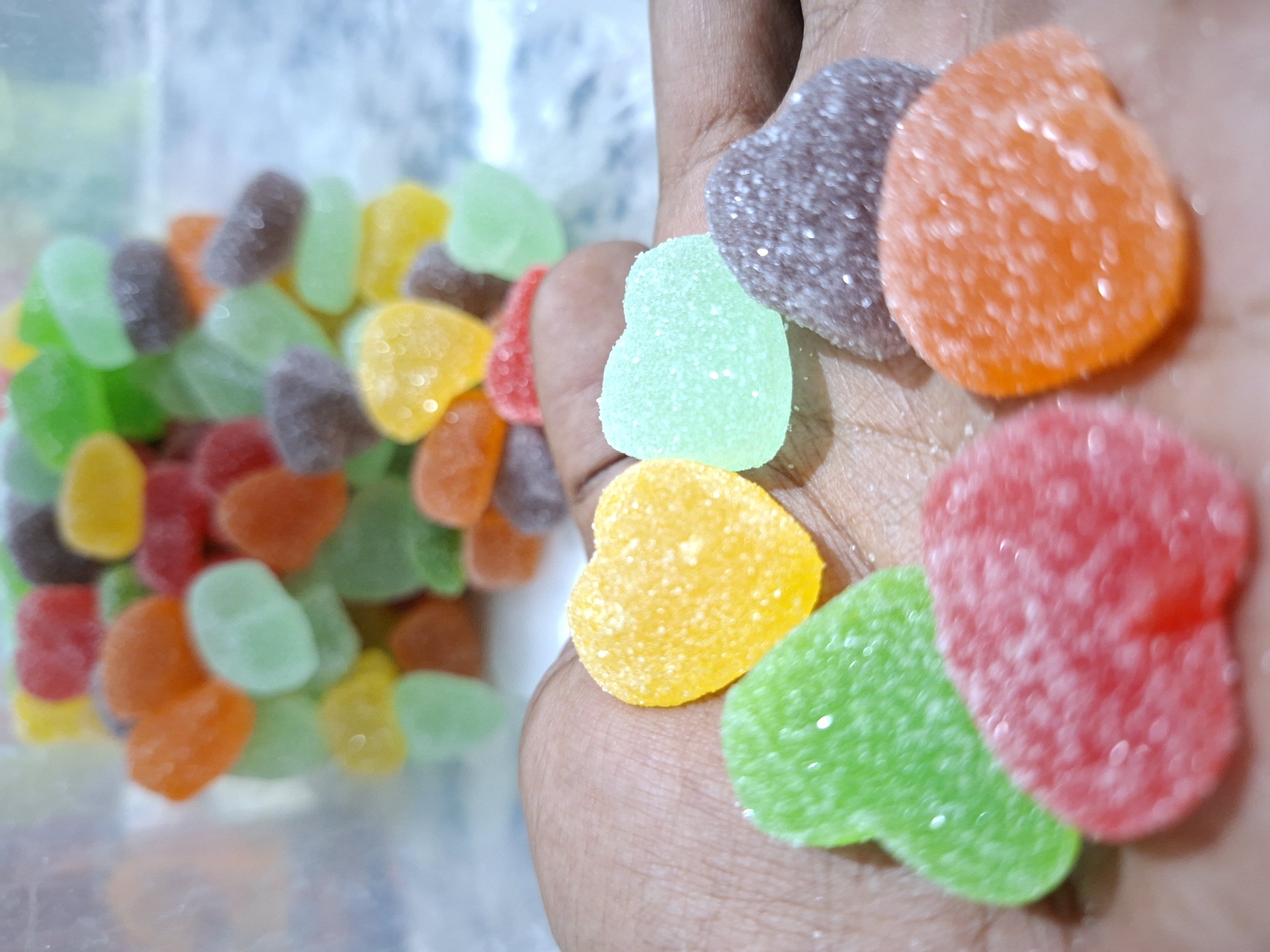 The image shows a person holding a handful of heart-shaped gummy candies of various colors. The candies appear to be coated in sugar. In the background, a clear container filled with similar candies is visible, along with a blurred, indistinct background possibly indicating an interior space. The specific location within Kutaglumpang, Indonesia, cannot be determined from the image.