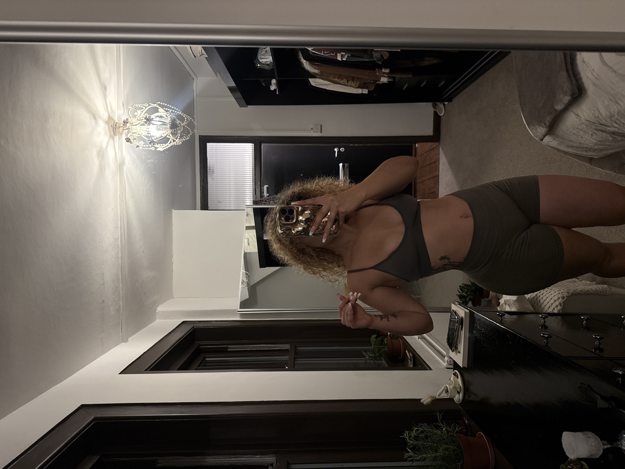 A person stands in front of a full-length mirror, taking a selfie. They are wearing a grey sports bra and olive green shorts. The person has curly blonde hair and a tattoo on their left arm. The mirror reflects a room with a dresser, a closet filled with clothes, a bed, and a chandelier hanging from the ceiling. The scene is likely a bedroom or dressing area.