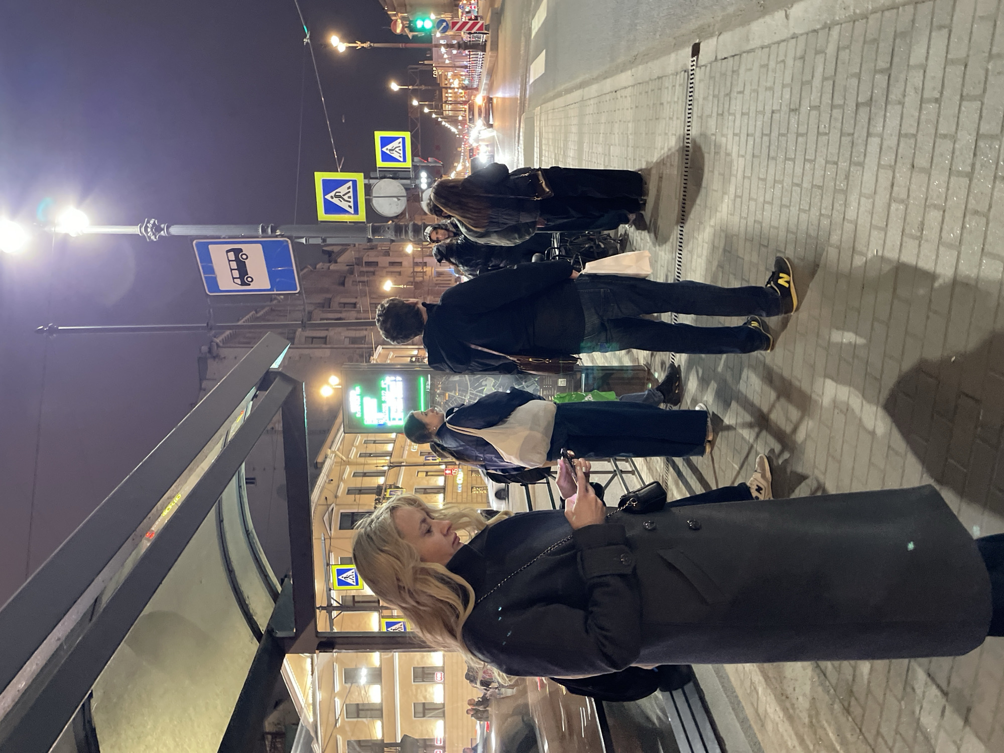 At night in Saint Petersburg, Russia, a bus stop shelters a blond woman looking at her phone. Other people are waiting on the sidewalk nearby. Pedestrian crossing signs are visible, and a bus stop sign is placed above the shelter. The street has moderate traffic, with lights from buildings and cars illuminating the scene.