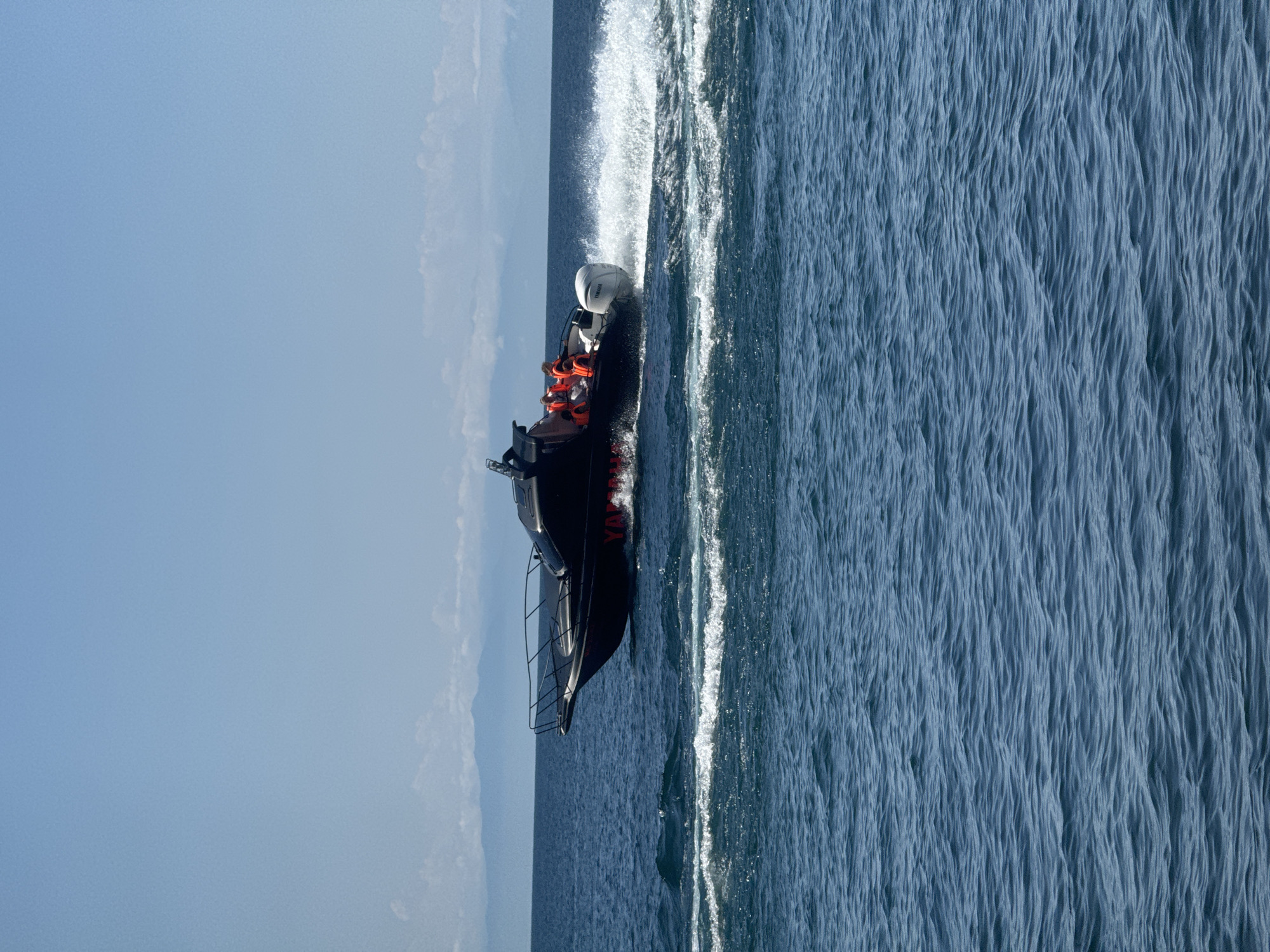 A black motorboat with an outboard engine travels across the deep blue water, creating a white spray in its wake. Several individuals wearing orange life vests are seated on the boat. In the background, a hazy mountain range under a clear, light blue sky can be seen. The scene is likely located on Lake Baikal near Listvyanka, Russia, given the typical landscape and the presence of boats.