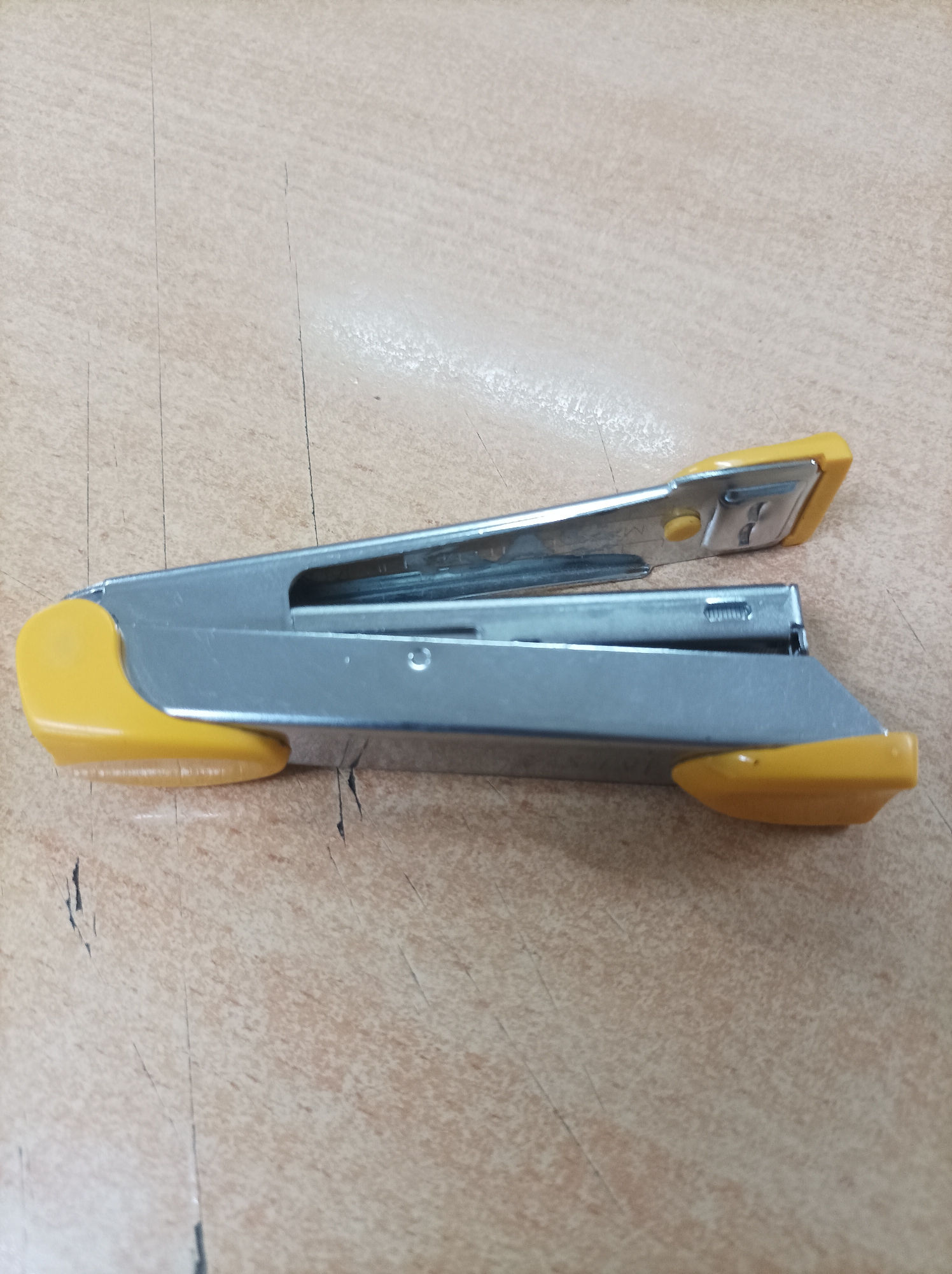 A silver and yellow stapler lies horizontally on a light brown, wood-grain patterned surface. The stapler is open, with the top arm slightly elevated. The surface appears to be a desk or table. There are no people visible in the image, and no actions are taking place. The lighting is adequate, highlighting the details of the stapler and the texture of the surface. The location is not specified within the image or any accompanying data.