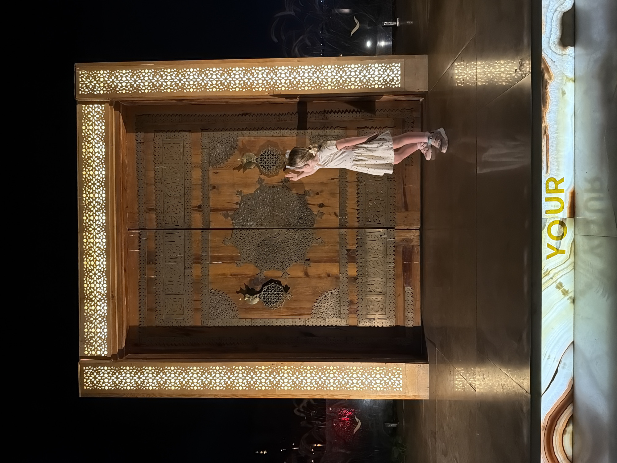 A young girl in a light-colored dress and shoes lies horizontally on a large, ornate wooden door, seemingly suspended in mid-air. The door is heavily decorated with metal patterns and borders, and it is framed with light-emitting panels. The area surrounding the door and the girl is dimly lit, but there are signs of light beyond in the background. A vertical, illuminated sign reading "YOUR" stands to the right of the door. This scene appears to be at a public display or attraction in Sharm El Sheikh, Egypt.