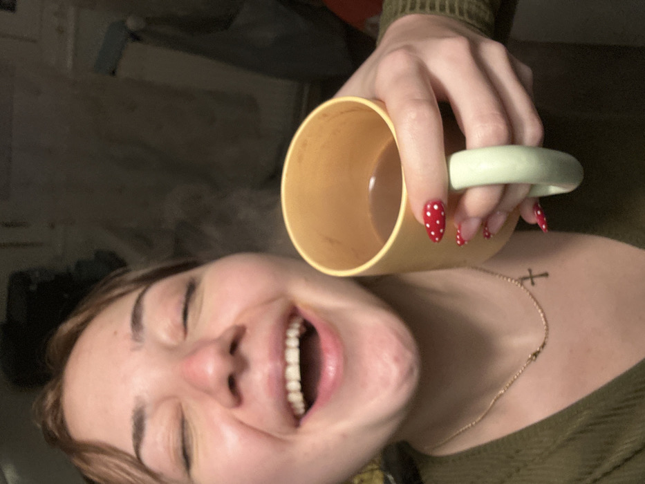 A woman smiles broadly, eyes closed, holding a yellow mug with a green handle near her face. Steam rises from the mug. She wears a green top and a gold necklace with a cross pendant. Her nails are red with white polka dots. The background is blurry and suggests an interior setting.
